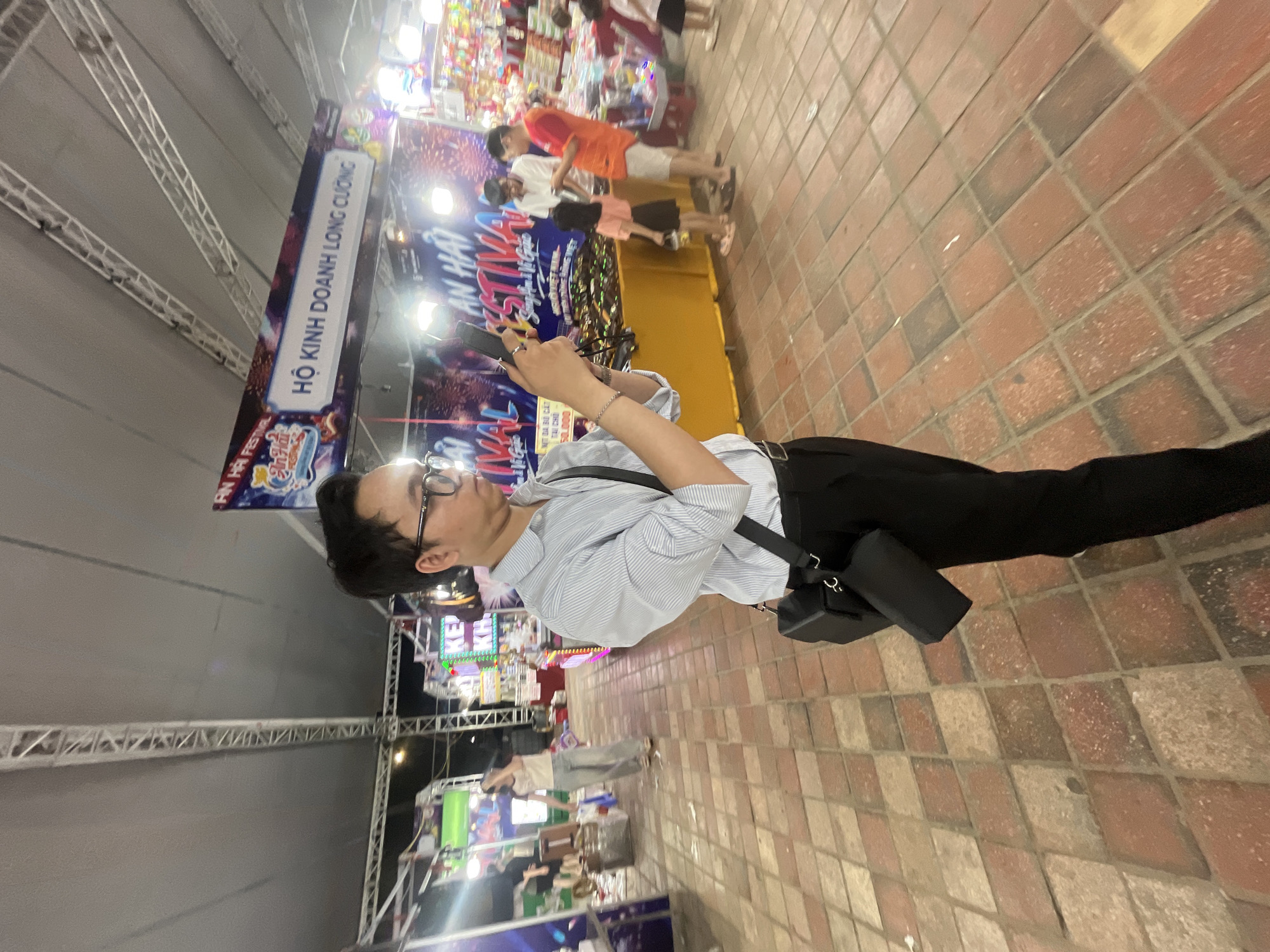 A young man in a light blue pinstripe shirt and black pants is standing outdoors at night, holding a smartphone and appearing to take a picture. He wears glasses and has a black crossbody bag. In the background, there are stalls with colorful decorations and signs, suggesting a festival or market. One sign above a stall reads "AN HAI FESTIVAL" and another says "HỘ KINH DOANH LONG CƯỜNG". The ground is paved with reddish-brown bricks arranged in a diamond pattern. Several other people, including a child, are visible in the background, interacting with the stalls. The scene is brightly lit, likely by artificial lights from the stalls and overhead coverings. This appears to be a commercial or recreational area, potentially part of the Anh Hai Festival in Da Nang, Vietnam, a coastal city known for its festivals and events.