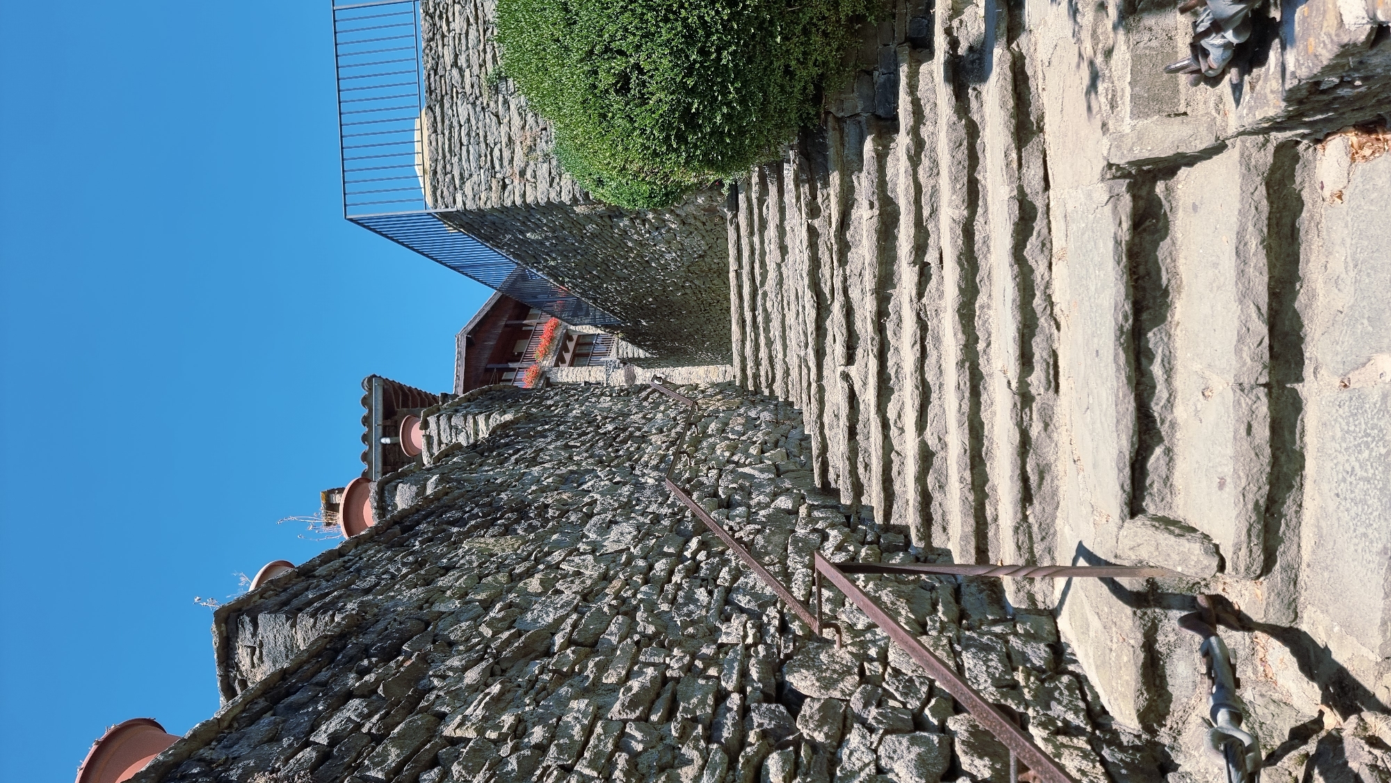 A stone staircase ascends between two stone walls under a clear blue sky in Rupit i Pruit, Spain. The left wall is constructed of rough, uneven stones and features a rusted metal handrail. The right wall is similarly constructed and is topped with a black metal railing, partially obscured by a dense green bush. At the top of the stairs, a wooden building with balconies adorned with red flowers is visible. Several large terracotta pots are placed on top of the walls. The scene appears to be a historic, outdoor location.