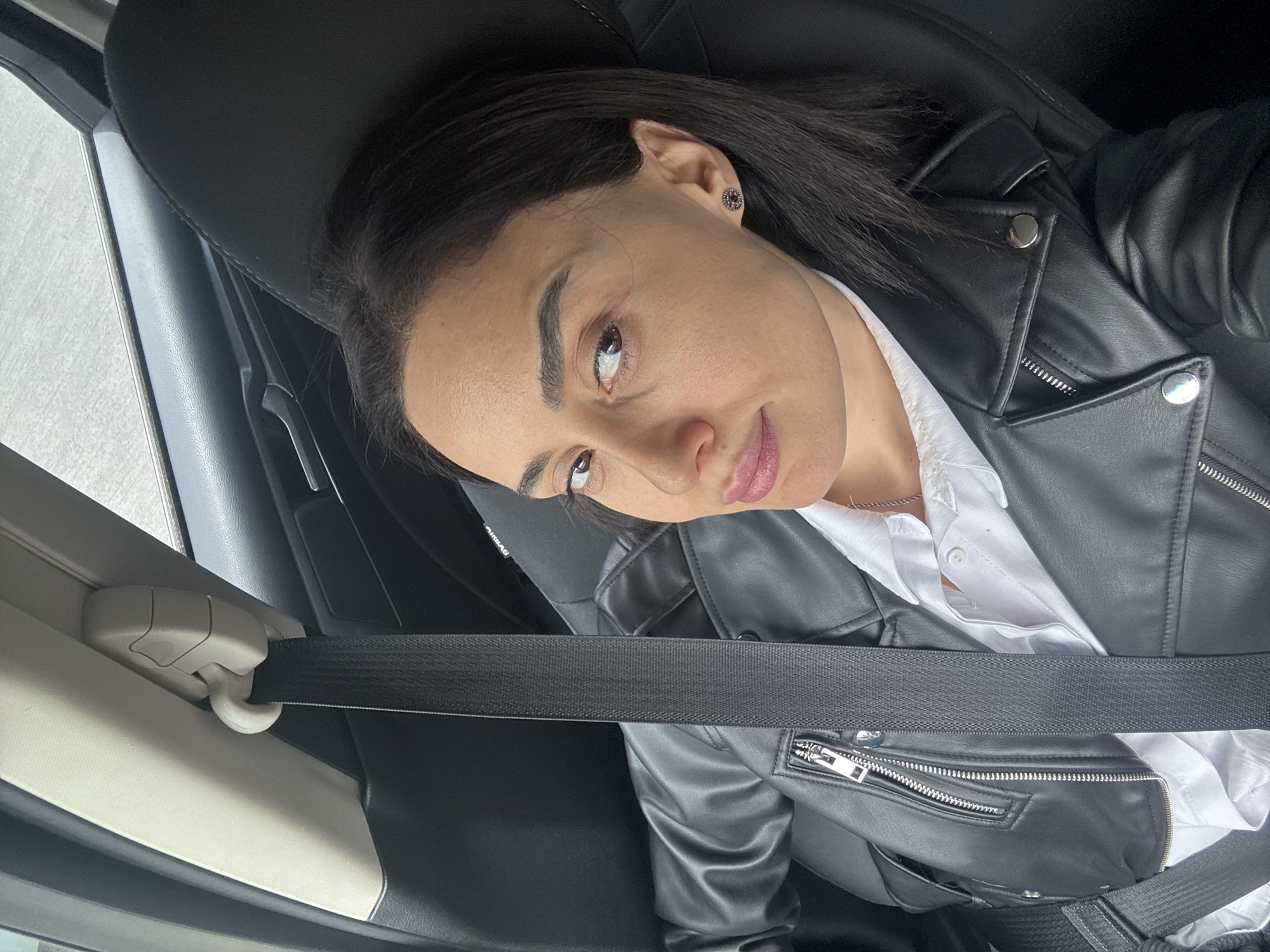 A woman is seated in a car, wearing a black leather jacket over a white collared shirt, and a seatbelt is fastened across her body. The location is Tbilisi, Georgia. The photograph is taken from a slightly overhead angle.