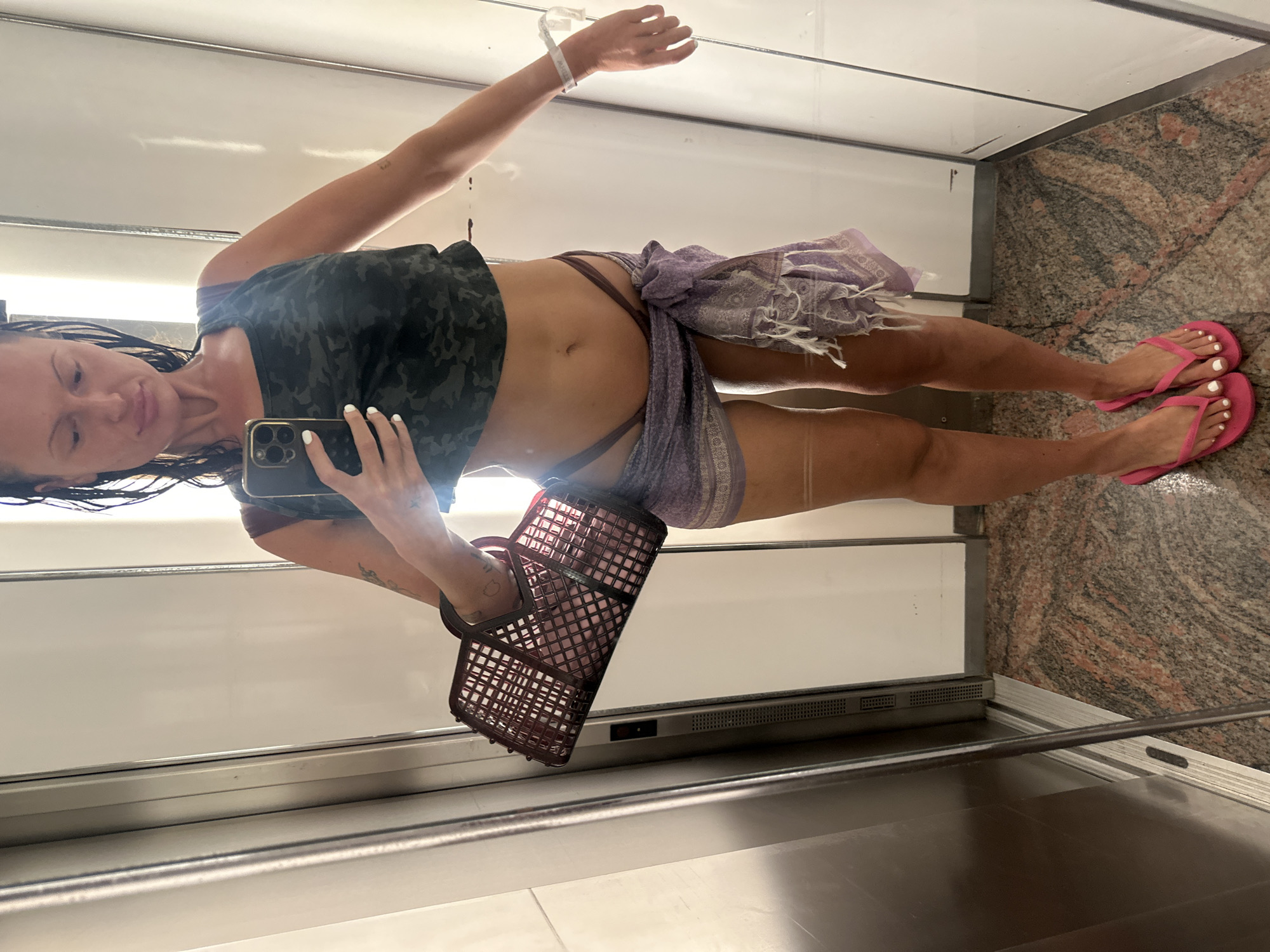 A full-body mirror selfie captures a woman standing in an elevator. The woman, with dark, wet hair, holds a dark smartphone in her right hand, facing her reflection to take the picture. She is dressed in a dark green camouflage crop top, dark brown bikini bottoms, and a light purple patterned sarong draped around her waist. A white wristband is visible on her left arm, which is raised. She carries a dark, rectangular, woven-style plastic bag on her left arm. Her feet are in bright pink flip-flops, and her toenails are painted white. Tattoos are visible on her right arm.
The elevator interior features light-colored, reflective walls that show distortions and reflections. The floor is composed of polished, variegated reddish-brown and gray granite or marble. A metallic strip runs along the base of the elevator walls. The setting is brightly lit, indicative of an indoor, likely daytime, environment. The wet hair, swimwear, and flip-flops suggest a casual, holiday context, potentially after swimming or showering. The woman's expression is neutral, as she focuses on the camera. No legible text is visible in the image.