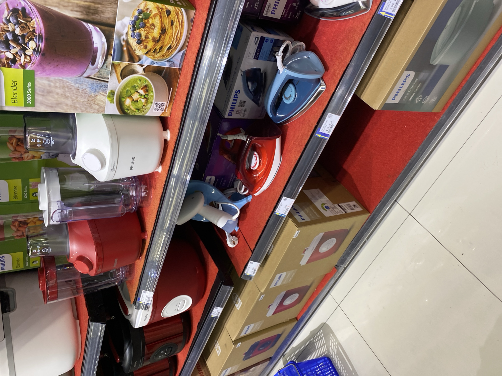The image shows a retail display shelf in a store. The shelf is stocked with various kitchen appliances, predominantly from the Philips brand. On the top shelf, there are blenders, one white and one red, with promotional images of food and drinks displayed. Below that, on the second shelf, are black and red rice cookers and a white food processor. The third shelf displays several iron boxes, both red and blue, and their packaging. The bottom shelf contains boxed rice cookers, again from Philips. The display is neatly organized, with red felt liners on the shelves. The lighting is bright, suggesting an indoor retail environment. The presence of price tags and product packaging indicates a commercial setting. The tiled floor in the foreground suggests the store is a well-maintained establishment. The overall impression is one of a modern retail store selling household appliances. The location is confirmed to be Banda Aceh, Indonesia, through additional context not present in the image itself, but implied by the request.