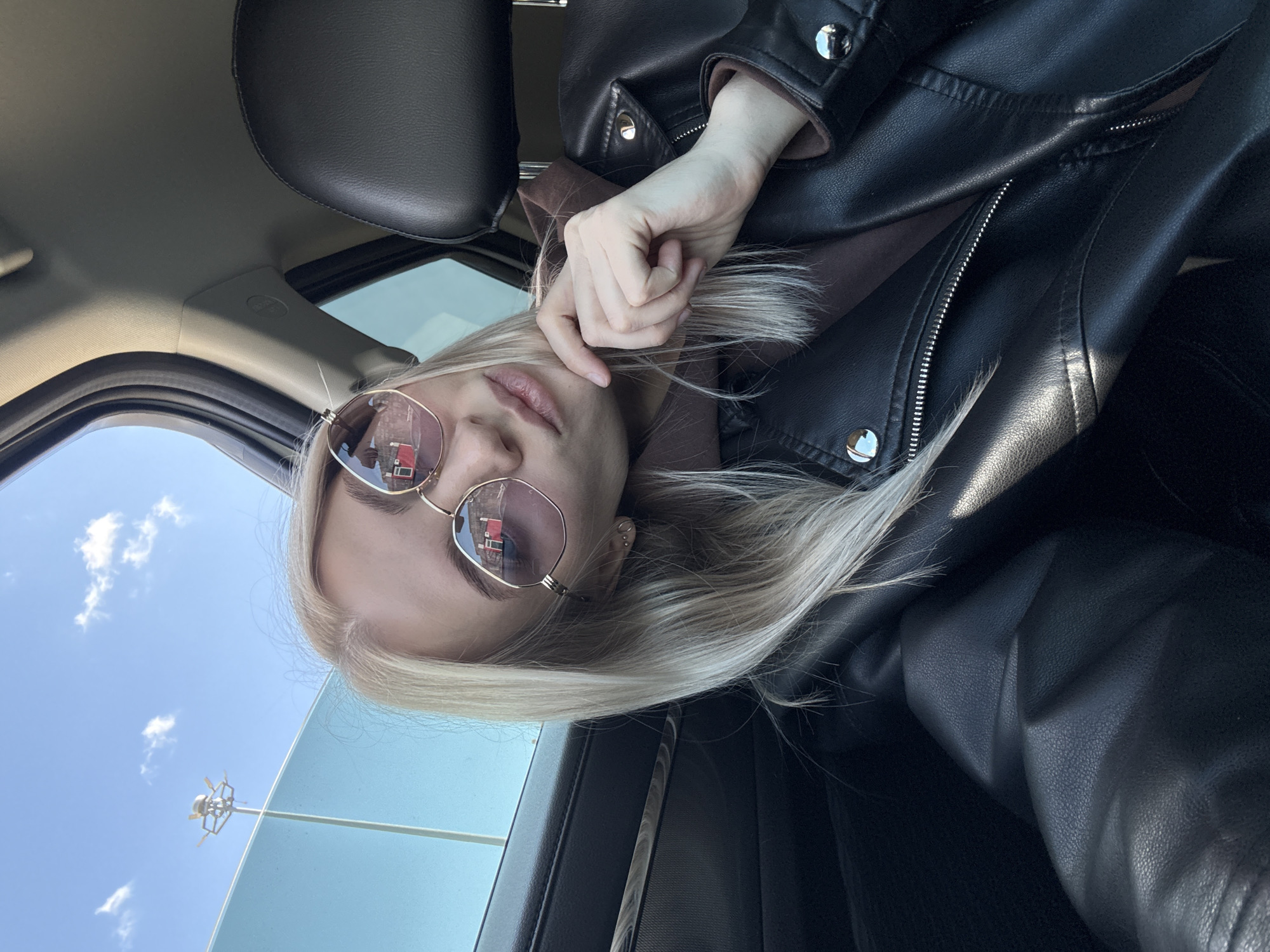 A woman with blonde hair, wearing rose-tinted sunglasses and a black leather jacket, is photographed in what appears to be the interior of a car. She is holding her hair with her right hand, positioning it near her face. Through the car window, a blue sky with scattered clouds can be seen, along with a light pole. The reflection in her sunglasses shows a red and white building. Considering the available visual information, the building's architecture, and the presence of urban infrastructure, it's possible that the reflection in the sunglasses could potentially be reflecting a building found within the city of Izhevsk, Russia, but this cannot be verified without the name of a specific building in Izhevsk, Russia that fits the details in the reflection. The interior of the car features black leather upholstery and a headrest. The angle of the photograph suggests it was taken either by the subject herself or by another person inside the vehicle.
