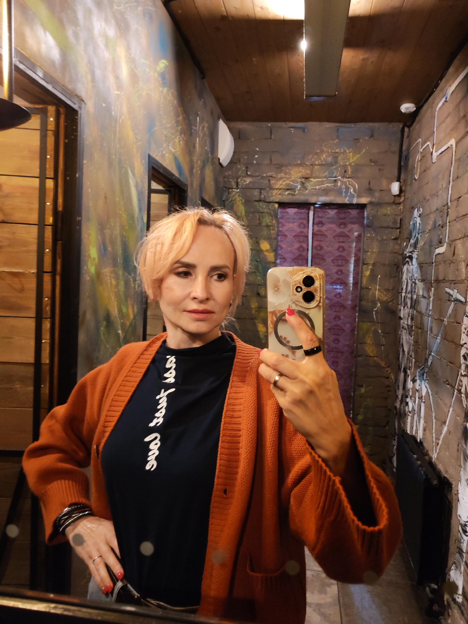 In a dimly lit interior space that appears to be a restroom or a narrow corridor with exposed brick walls adorned with graffiti, a blonde woman with short, peach-colored hair takes a mirror selfie. She is wearing a dark blue t-shirt with white cursive writing and an open, rust-colored cardigan. She holds a smartphone with a floral case in her right hand, displaying the camera's view. The background reveals wooden paneling on the left, a dark purple door with a patterned design, and a radiator on the right. The overall ambiance is urban and somewhat gritty, suggesting a public venue like a bar or club. The city of Bol'shie Mkhi, Russia, is mentioned as the location, though the visual cues do not provide specific indicators of this particular locale, relying more on general urban aesthetics.