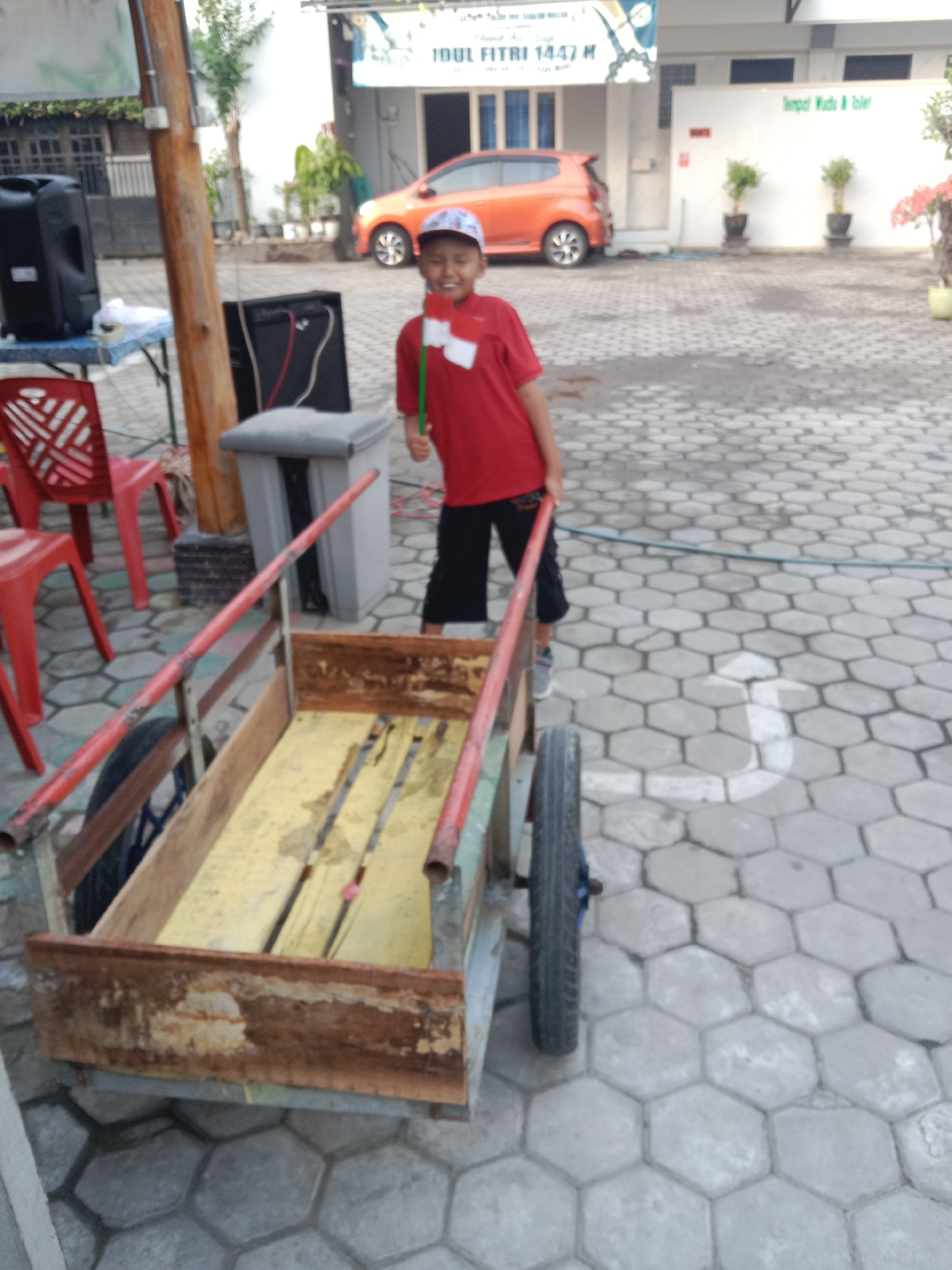 A young boy in a red shirt and a white and blue baseball cap is standing next to a wooden cart on a paved ground area. He is holding a small Indonesian flag. The cart has red handles and a single wheel. In the background, there is an orange car, a building with a banner that reads "IDUL FITRI 1447 M", and some potted plants. The paving stones are hexagonal. Red chairs are visible to the left of the boy and the cart. A speaker is placed on a blue table near the chairs. A wooden pole stands to the left of the boy. There is a trash can behind the boy. The scene appears to be outdoors, possibly in front of a building or in a public space. The white and blue banner suggests the image was taken around Eid al-Fitr, a Muslim holiday. The mention of "Tempat Wudlu & Toilet" on the building indicates a place for ablution and restrooms.