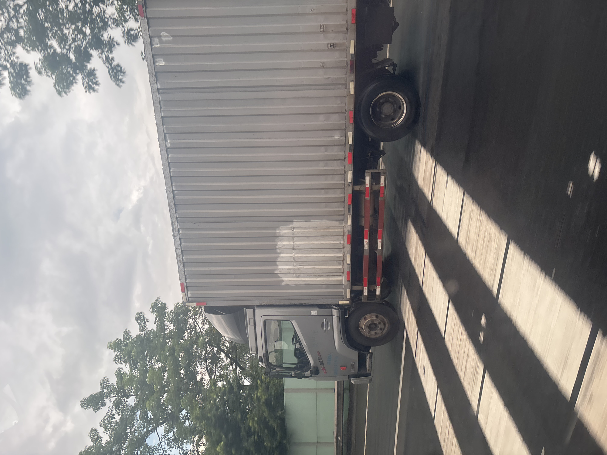 A silver box truck, with a ribbed metal cargo container, is driving on a multi-lane asphalt road. The truck has a grey cab with a black roof-mounted spoiler. Red and white reflective markers are visible along the side of the cargo container. The truck is in motion, as indicated by the motion blur on the wheels and the road. Sunlight casts diagonal shadows across the road, creating a striped pattern. Green trees and foliage are visible on the left side of the frame, bordering the road. The sky is cloudy with patches of blue. The scene appears to be in an urban or suburban area, possibly along a highway in Baiyun District, China.