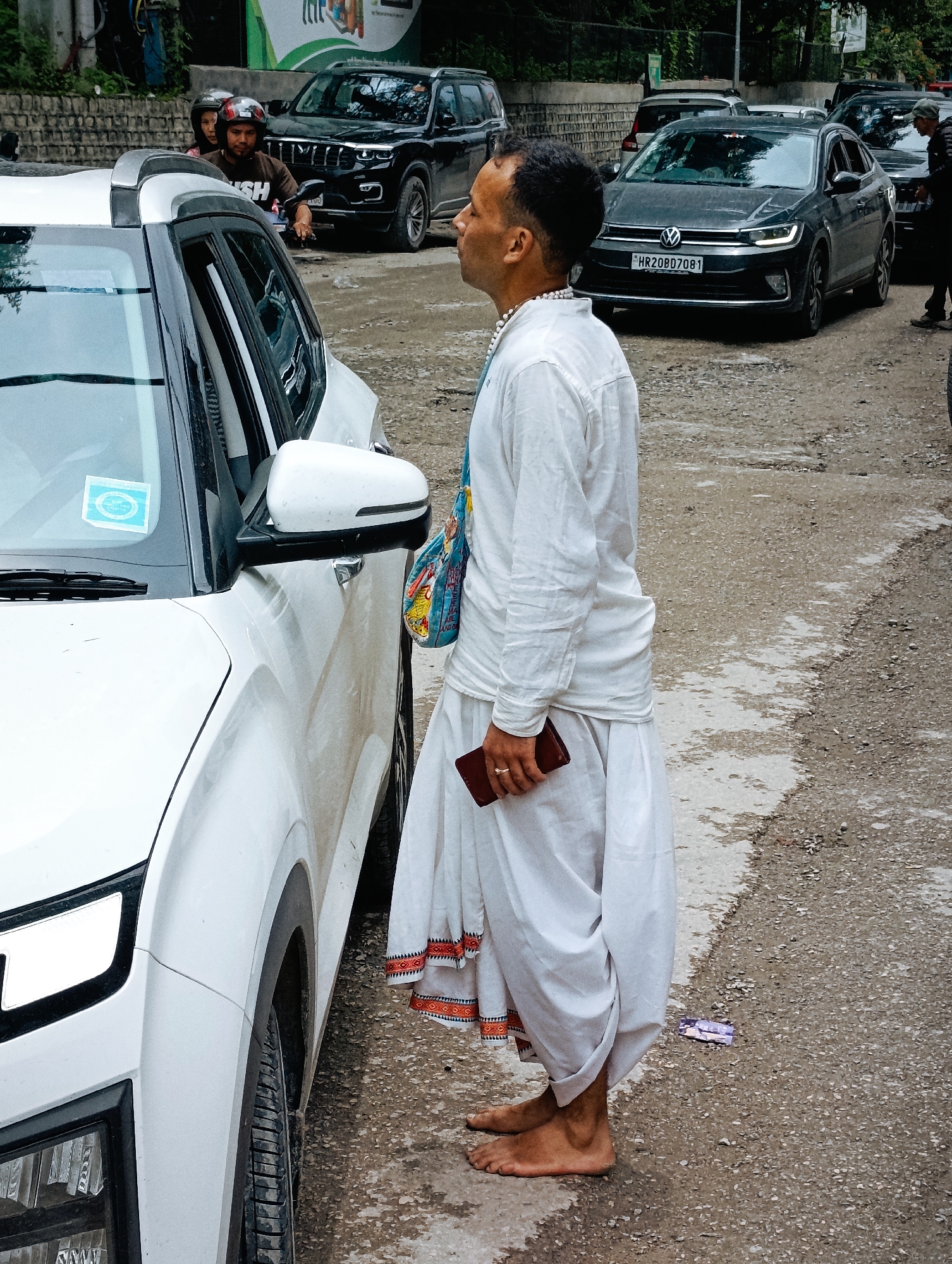 The image captures a street scene in Manali, India, likely on a cloudy day given the diffused lighting. In the foreground, a white SUV is parked, its right side prominently featured. A man, dressed in traditional white attire with a dhoti-like lower garment and a long-sleeved shirt, stands next to the SUV. He is holding a dark-colored mobile phone in his right hand and appears to be looking away from the camera, towards the right side of the frame. He wears a necklace of white beads. His bare feet are visible on the rough, paved ground, which appears damp in places.
Behind the white SUV, other vehicles are visible on the street, including a dark-colored SUV and a black Volkswagen sedan with license plate HR20BD7081. In the background, there are hints of trees and buildings, consistent with an urban or semi-urban environment in Manali. A couple on a motorcycle, wearing helmets, are visible in the far background, behind the white SUV. The overall atmosphere suggests a normal day on a street in Manali with pedestrian and vehicle traffic.