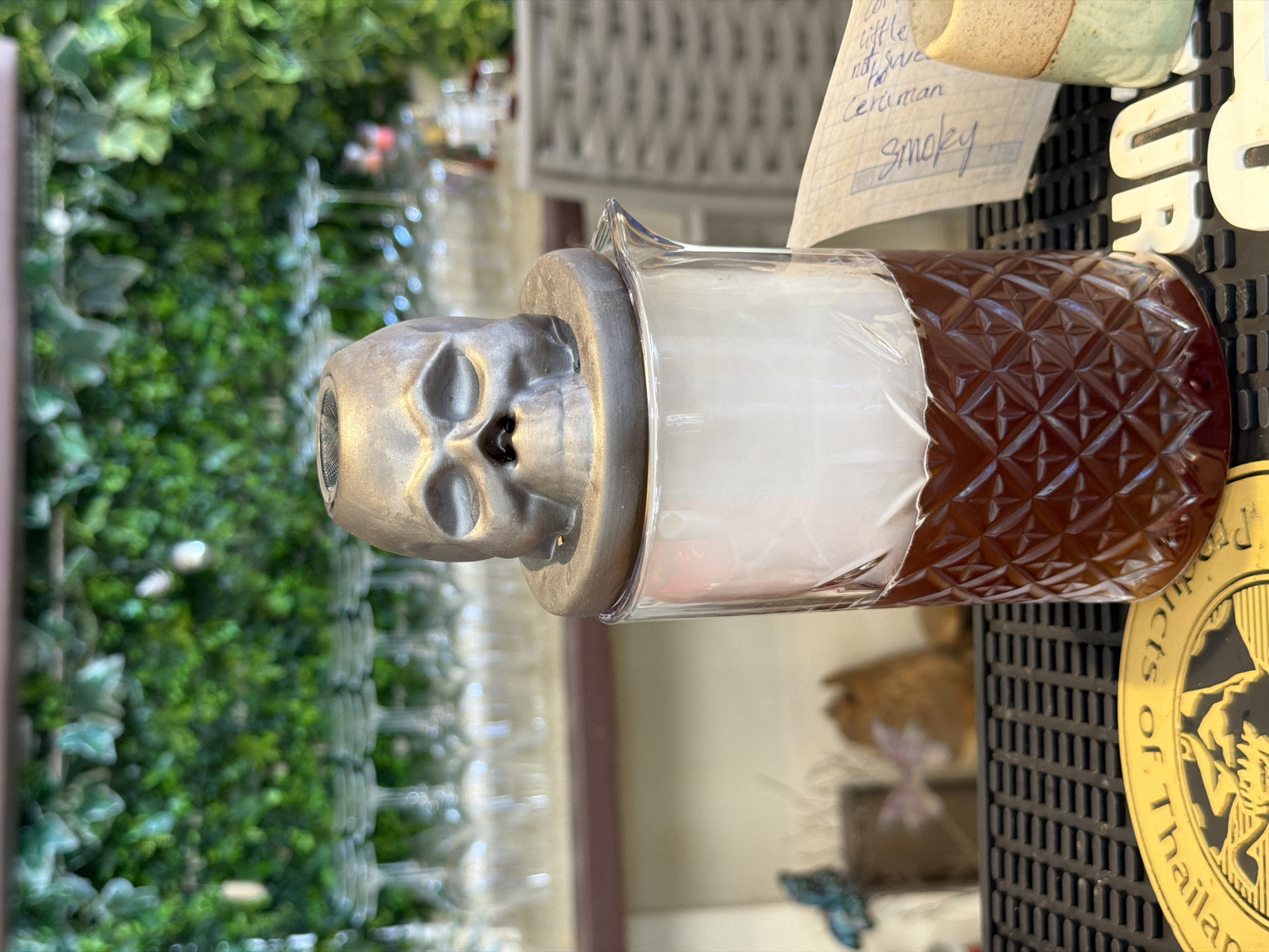 A close-up, eye-level, outdoor shot captures a unique cocktail presentation at a bar in Baan Thong Krut, Thailand. The foreground is dominated by a dark ribbed bar mat. Resting on the mat is a patterned glass carafe filled with a dark amber liquid, topped with a thick layer of white smoke. A silver, skull-shaped lid rests on top of the carafe, obscuring the top of the glass. To the right of the carafe, a piece of paper with handwritten notes, including the word "Smokey," is visible. A small ceramic cup with a speckled glaze sits partially in the frame to the far right. In the background, rows of clean glassware are arranged on a shelf. Behind the glassware, a lush green wall of artificial foliage provides a vibrant backdrop. The overall lighting suggests it is daytime.