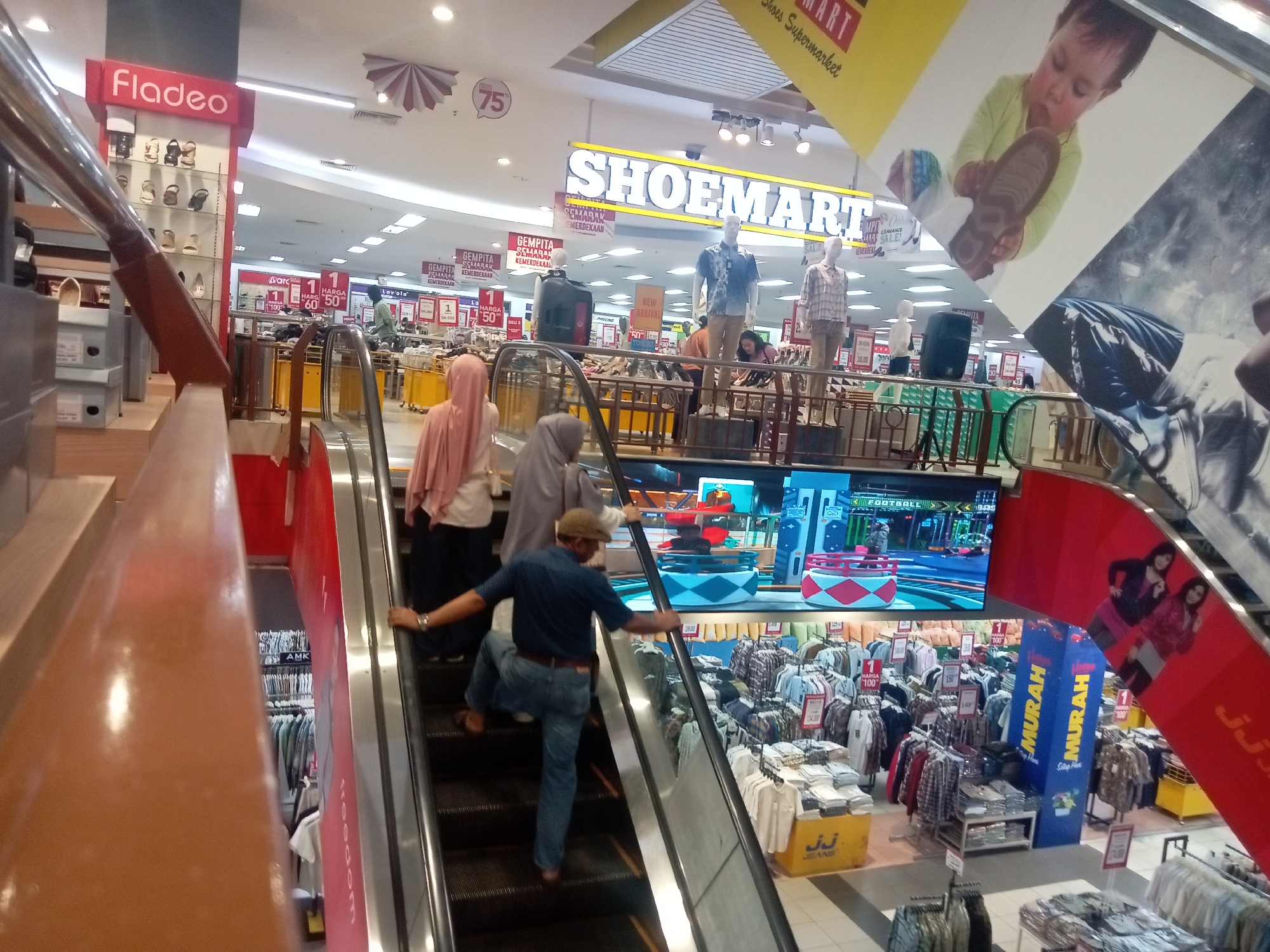 The image depicts the interior of a large department store, likely in Batam, Indonesia, featuring multiple levels and escalators. A prominent sign reads "SHOE MART," indicating a focus on footwear. Mannequins are displayed wearing casual clothing, and racks of shirts and apparel are visible throughout the store, suggesting a wide selection of merchandise. Several individuals are present, including shoppers ascending an escalator, some wearing headscarves and casual attire. The environment is brightly lit with fluorescent ceiling lights and illuminated signage. A large screen in the background displays a video game or promotional content. The overall impression is one of a busy, modern retail environment.