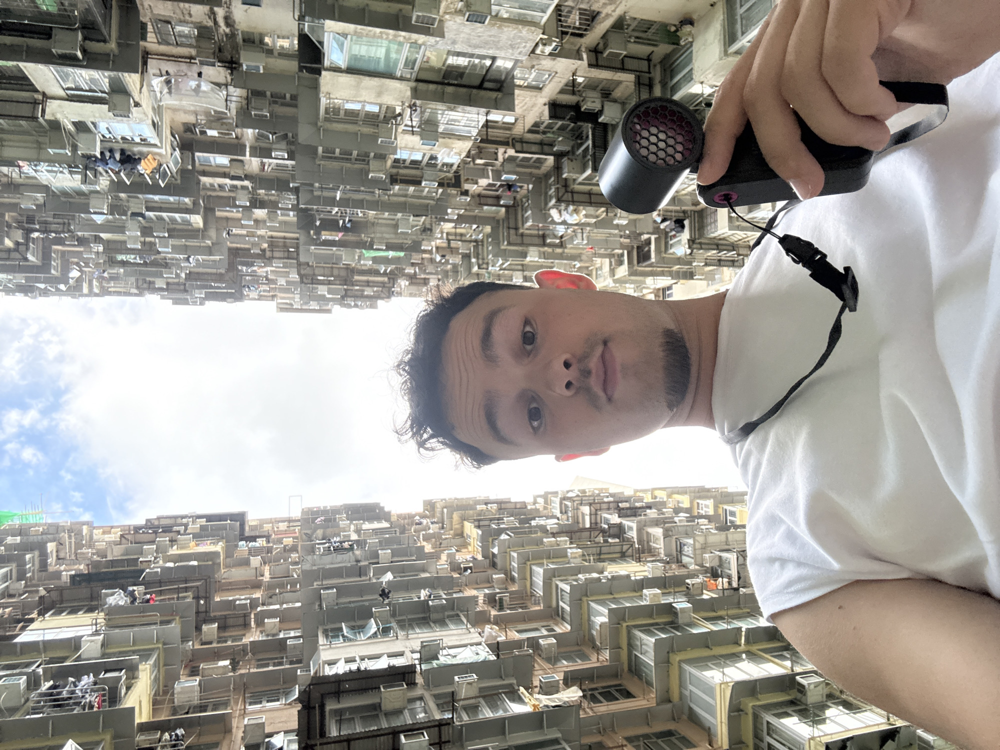 A person in a white t-shirt is holding a small, black device with a lanyard attached. The background reveals a densely populated urban area in Hong Kong, China. The architecture consists of numerous apartment buildings tightly packed together, many with small windows and external air conditioning units. Some laundry can be seen hanging outside the windows. The sky is partly cloudy. The image is upside down.