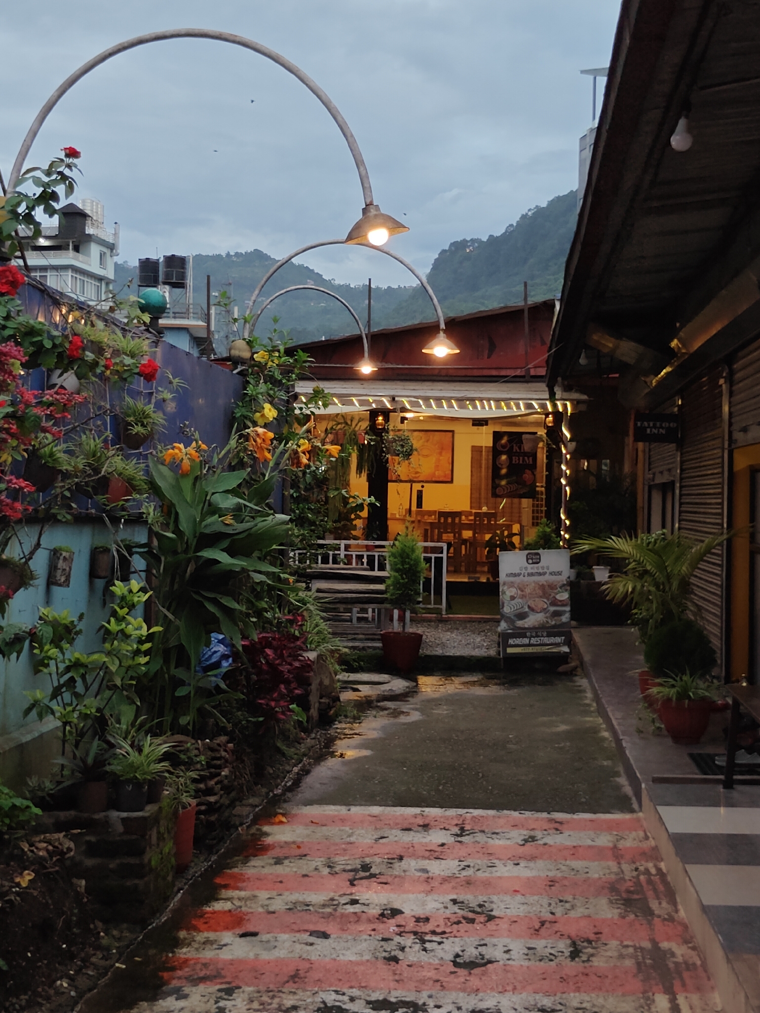 The image captures a view of a pathway leading into a restaurant establishment, likely in Pokhara, Nepal, given the presence of lush greenery and a mountainous backdrop. The pathway is paved with stones and has red and white stripes. On the left, a wall is adorned with various potted plants and flowering vines. The pathway leads to an open-air seating area of a restaurant with tables and chairs visible. Further back, the restaurant interior is illuminated, showcasing artwork on the walls and bamboo accents. A sign indicates "Kimbap & Bibimbap House" and "Korean Restaurant". To the right of the pathway, there's a building with a wooden facade, featuring a sign for "TATTOO INN". The sky is overcast, suggesting it might be dusk or a cloudy day. Several arched structures with lights are present overhead, creating an inviting ambiance. The overall scene depicts a commercial area with a blend of natural elements and urban infrastructure.