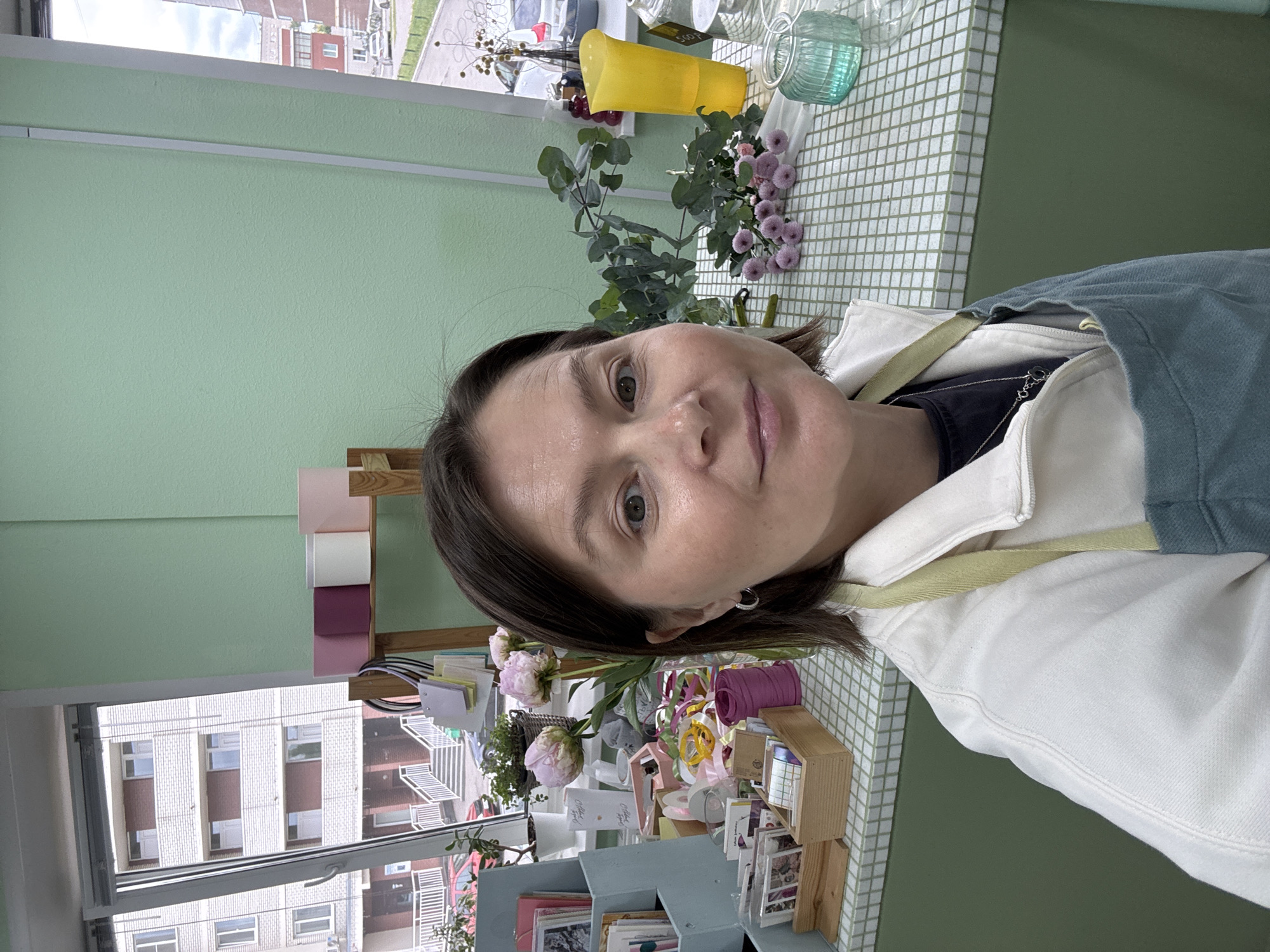 A woman is posing for a selfie indoors. She has brown hair and fair skin. She's wearing a light-colored jacket over a dark shirt with a light blue apron. Behind her is a light green wall and a window showing a multi-story building. On a counter are various items, including flowers in pots and vases, ribbons, cards, and office supplies. Some buildings are visible through the window. The location is identified as Izhevsk, Russia.