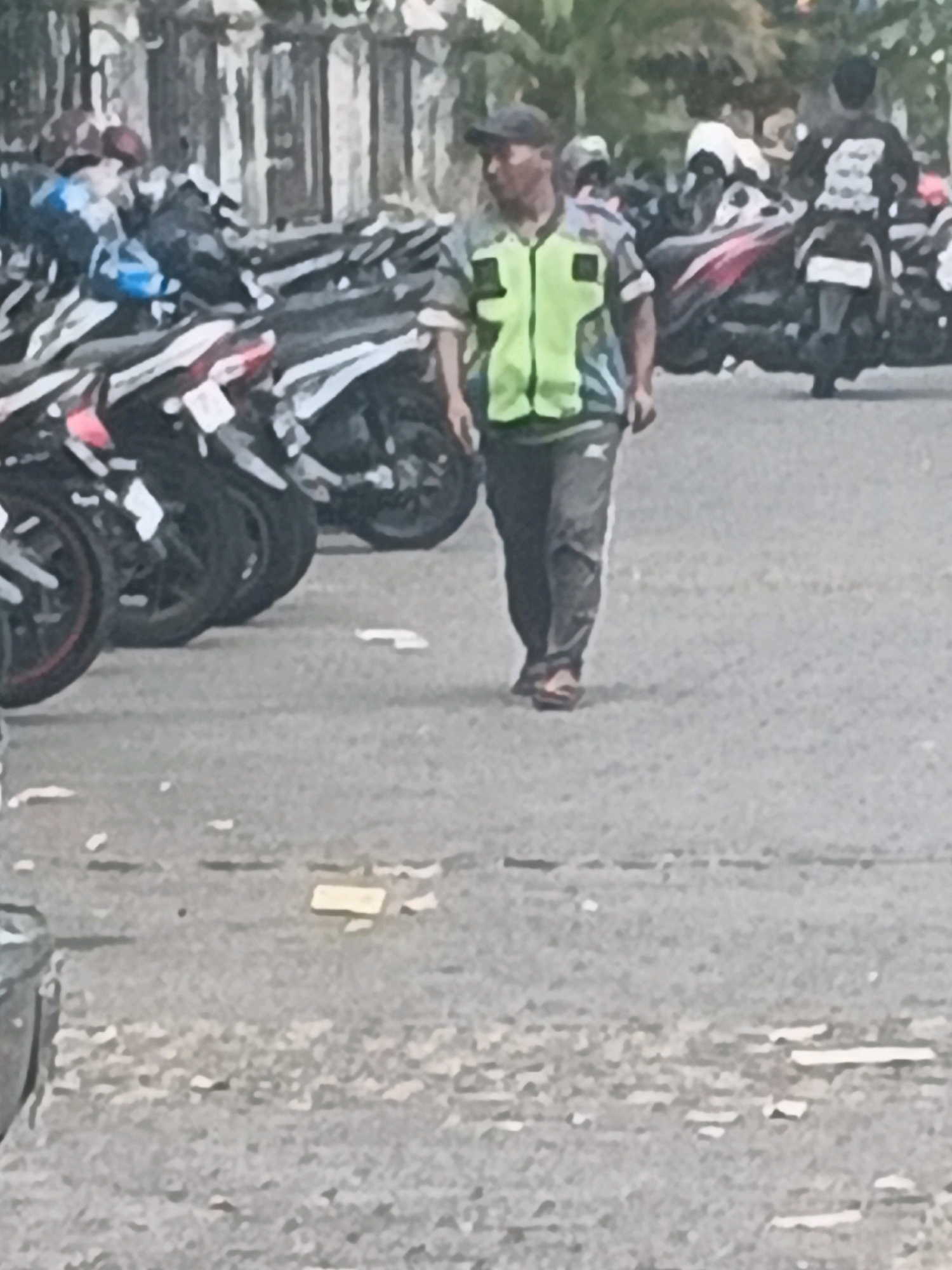 In Kalirejo, Indonesia, a man in a bright green and black vest walks past a row of parked motorcycles. The man wears a dark cap and dark pants. Several motorcycles are visible, parked side-by-side. The ground is paved and appears to be a parking area. The background shows blurred buildings and some vegetation. The scene is brightly lit, suggesting daytime.
