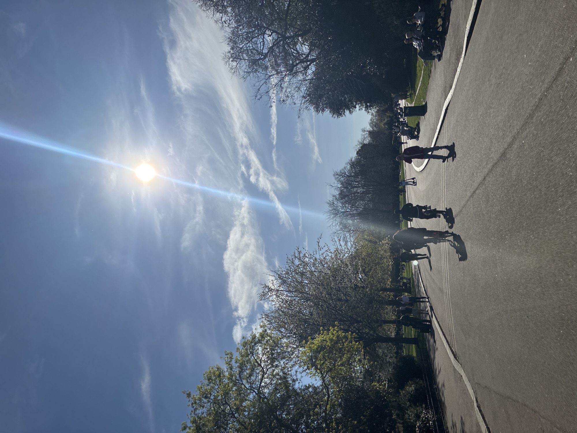 A horizontally oriented image depicts a sunlit park pathway in London, United Kingdom. The sun shines brightly in the blue sky, streaked with wispy clouds. Trees line the path, their leaves partially obscuring the sunlight. On the pathway, several people can be seen walking and cycling. Some people are seated on benches along the path. The pathway is dark gray and is separated from the green grass by a white line.