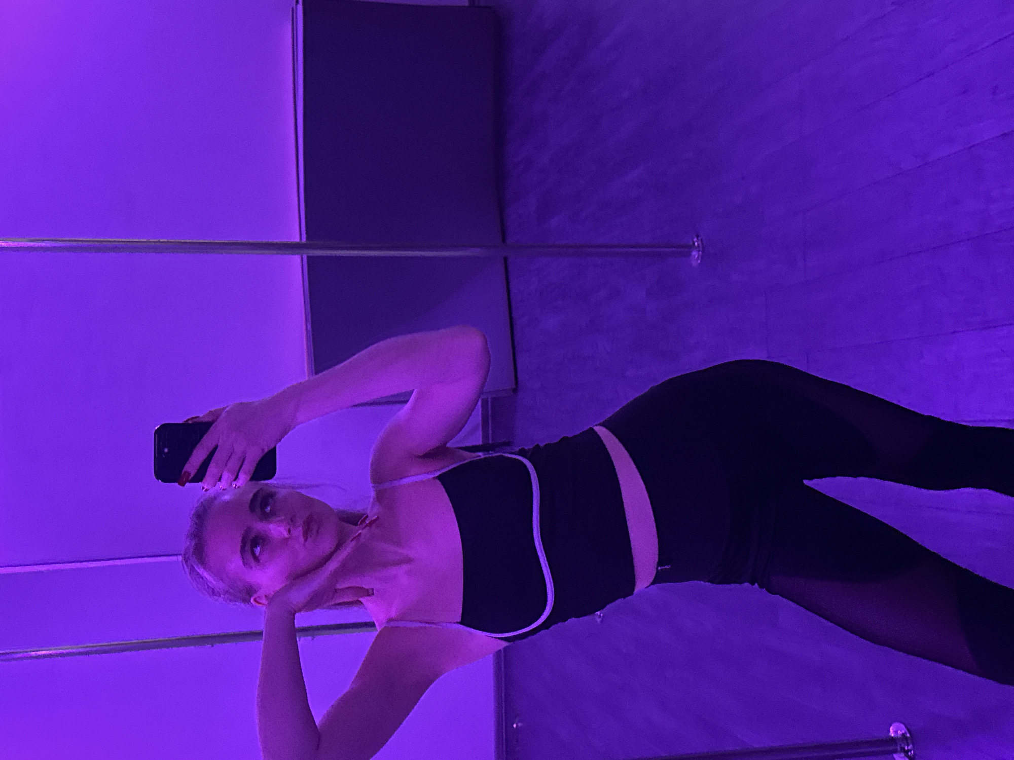 A young woman is taking a selfie in an Unknown, Unknown dance studio. She is wearing a black athletic top with white trim and black leggings with sheer panels. She is holding a black smartphone in her right hand, raised above her head. Her left hand is positioned near her face. A silver dance pole is visible in the background, extending from floor to ceiling. The studio is illuminated with purple lighting, highlighting a parquet wood floor and dark gray walls.