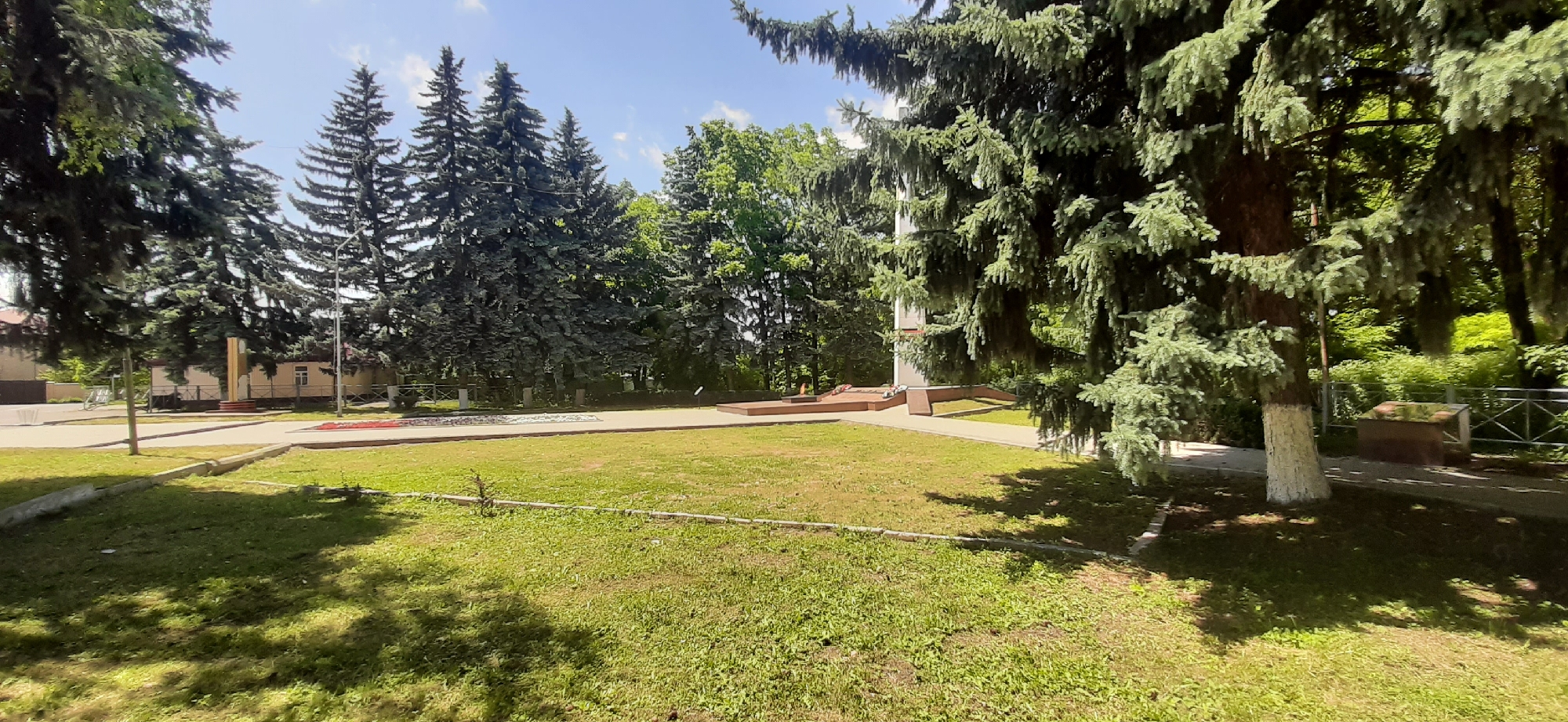 The image depicts a park area in the city of Yessentukskaya, Russia, on a bright, sunny day. In the foreground, a grassy lawn is dappled with sunlight and shadow from the surrounding trees. A low, concrete curb runs along the edge of the lawn. In the midground, a paved pathway leads towards a memorial. The memorial consists of a tall, slender obelisk or monument, with a base and surrounding plaza constructed of reddish-brown stone. There are some floral arrangements visible at the base of the memorial, and what appears to be an eternal flame, a common feature in war memorials. Behind the memorial, a row of tall evergreen trees stands against a bright blue sky with scattered white clouds. To the left of the memorial, several more evergreen trees are visible, along with a low, single-story building in the distance. To the far left, a light pole with a single lamp is present. The right side of the image shows more dense foliage and a metal fence. The overall scene is one of a public park with a significant monument, suggesting a place of remembrance or historical importance. There are no people or animals visible in the image.