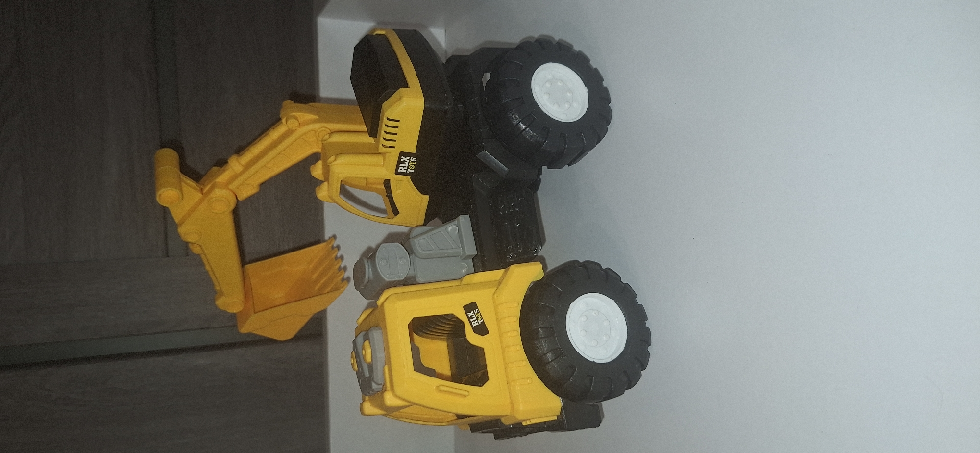 A yellow toy excavator is displayed on a white surface, with a wooden-paneled wall in the background. The excavator has a black chassis and yellow cabin and shovel arm. The cabin features "RLX TOYS" stickers on both sides. The excavator has large, black, treaded tires with white rims. The background wall has vertical lines, suggesting a wood grain or paneling. There is no indication of the location being Bila Tserkva, Ukraine, or any specific city.