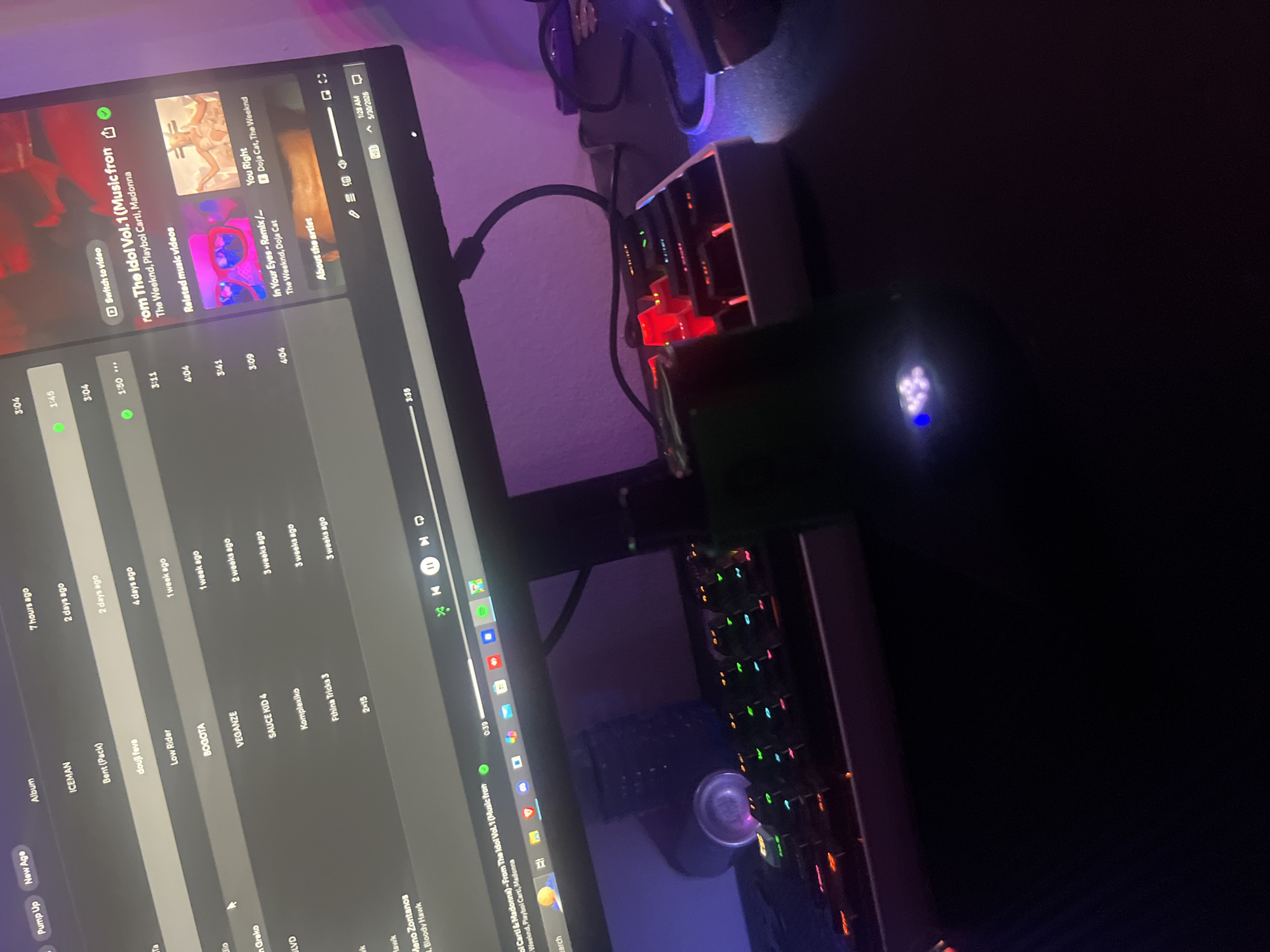 The image shows a dimly lit workspace with a computer monitor displaying Spotify. The monitor screen displays music from "The Idol Vol. 1 (Music fron" by The Weeknd, Playboi Carti, and Madonna. Nearby, there is a mechanical keyboard with colorful backlighting and a dark, cylindrical object with a blue illuminated button on top. The image does not provide sufficient information to determine a specific location within the city of Paphos Municipality, Cyprus.