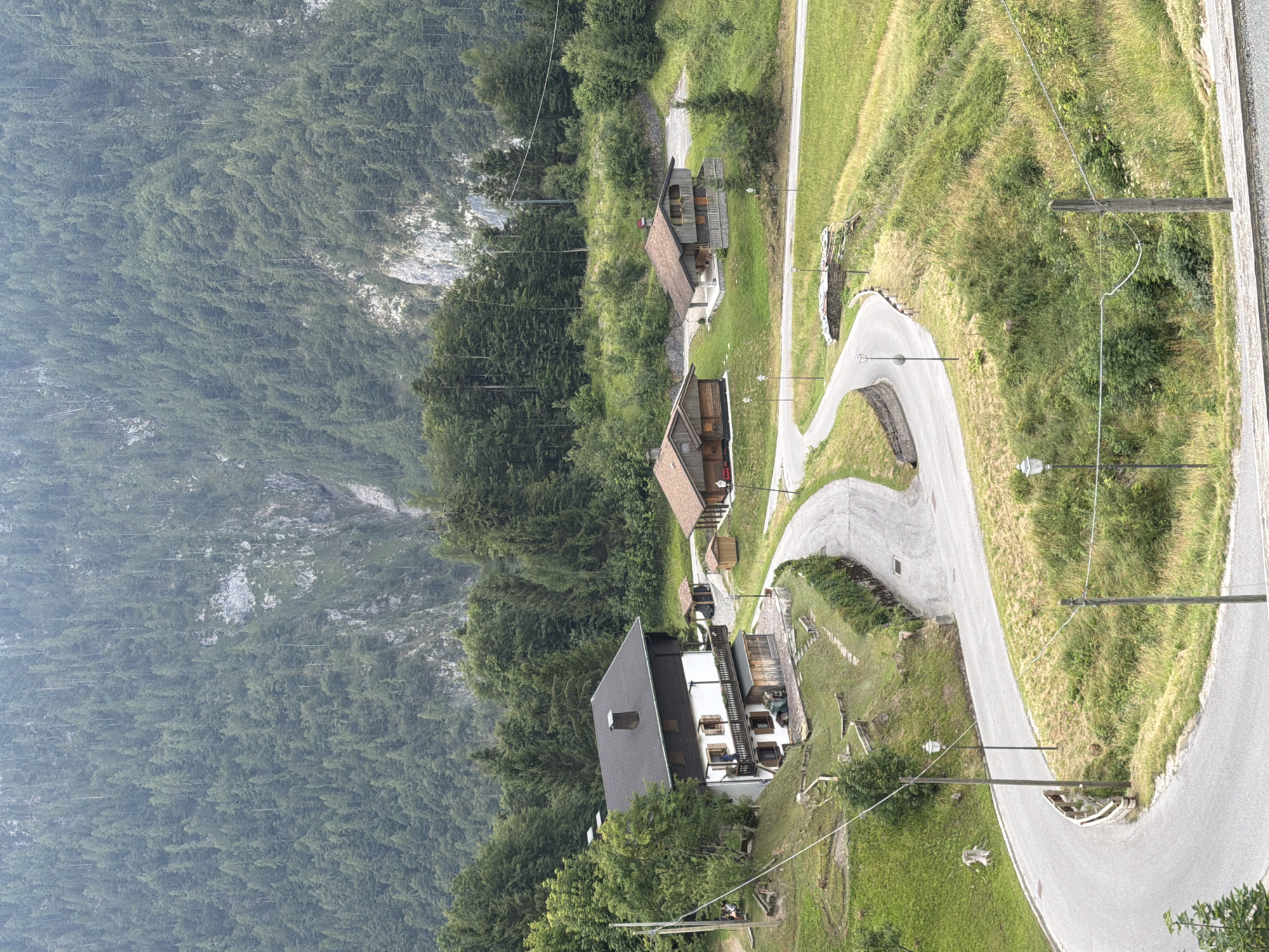 The image depicts a mountainous landscape in Sauris / Zahre, Italy, with a winding road leading past several traditional wooden houses and a larger white house with a dark roof. The houses are nestled on grassy slopes dotted with trees, and a dense forest covers the steep mountainside in the background. Several streetlights line the road. In the foreground, a group of people are seated at picnic tables near the white house.