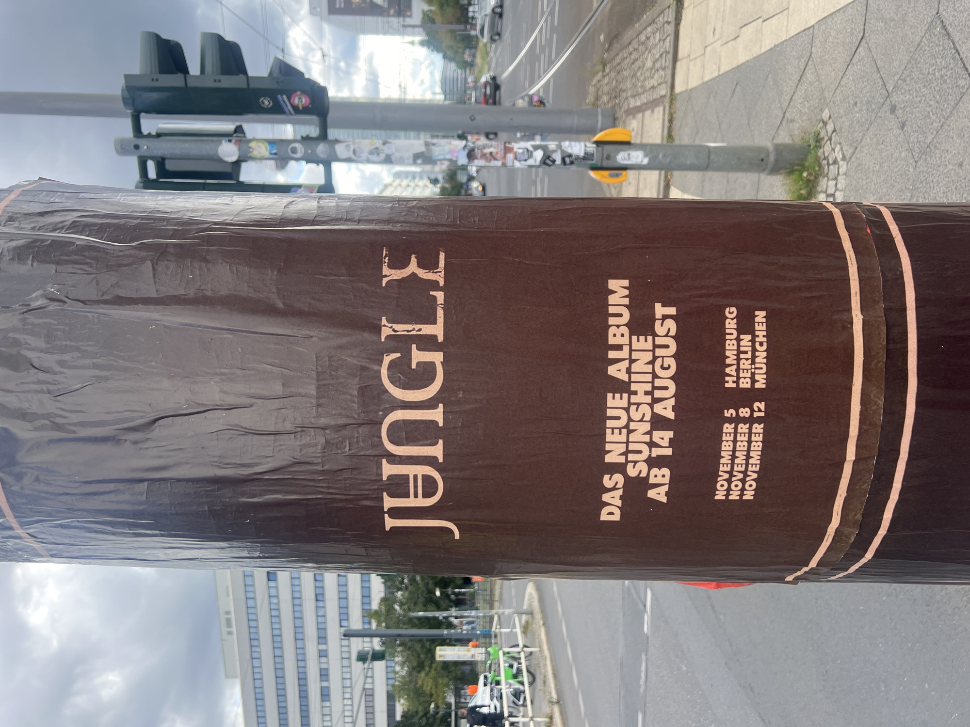 The image shows a brown poster wrapped around a lamppost on a city street. The poster features text and graphics related to an album release. The text "JUNGLE" is prominently displayed in large, stylized letters at the top. Below this, it reads "DAS NEUE ALBUM SUNSHINE AB 14 AUGUST". Further down, there is a list of dates and cities: "NOVEMBER 5 HAMBURG", "NOVEMBER 8 BERLIN", and "NOVEMBER 12 MÜNCHEN".
The lamppost is metallic gray and has a traffic light attached to it. The background shows a street with cars and trams, buildings, and pedestrians, suggesting an urban environment. The overall impression is that of an outdoor advertisement in a public space.
Based on the information on the poster, the location is likely Berlin, Germany, as one of the listed cities is "BERLIN". The poster announces an album called "SUNSHINE" by "JUNGLE" and lists concert dates in Hamburg, Berlin, and Munich in November. The album release date is given as August 14th. The image appears to have been taken during the day, with cloudy skies.