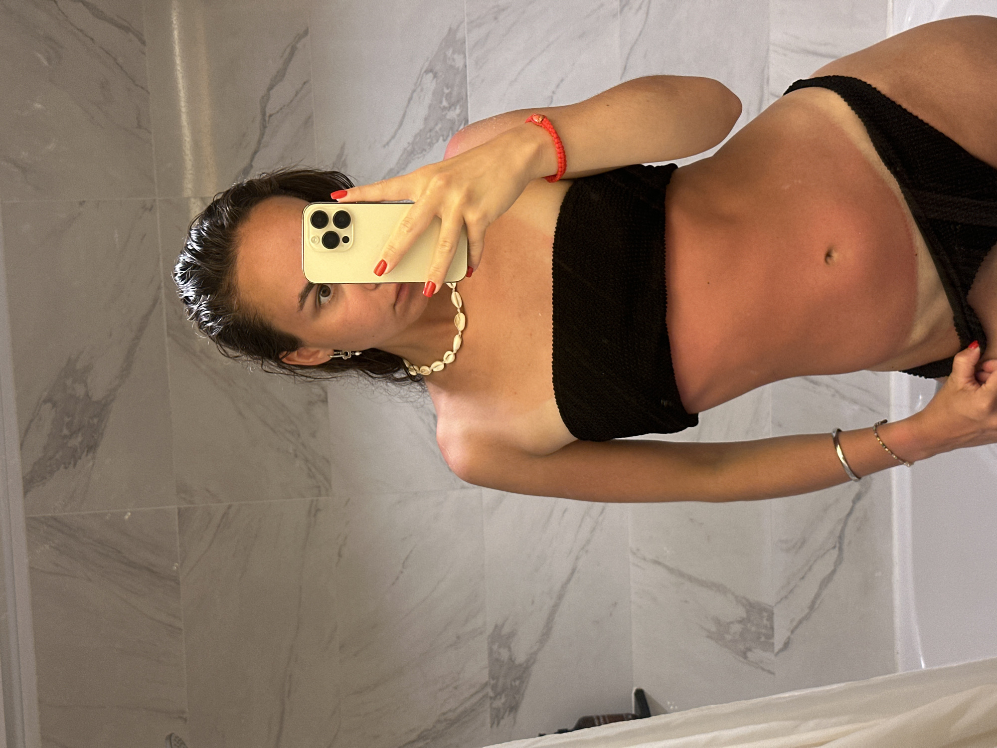 A tanned woman in a black two-piece swimsuit takes a mirror selfie with her phone in a bathroom. The woman has wet, dark hair, fair skin, and is wearing a red bracelet, a seashell necklace, and two bracelets on her right wrist. The bathroom has marble-patterned walls and a white tub. It is impossible to verify the location.