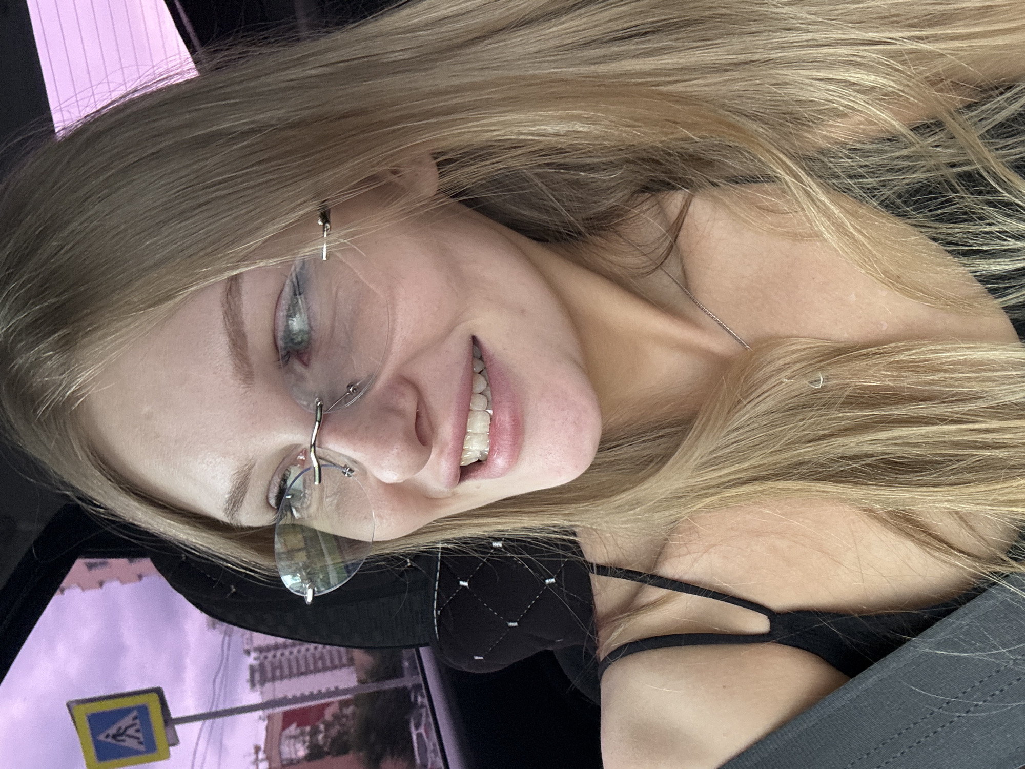 The image shows a close-up, indoor shot of a young woman with long, blonde hair, wearing rimless glasses and a black tank top. She is seated in a car, evidenced by the seatbelt visible in the foreground and the tinted window in the background. Outside the window, a pedestrian crossing sign is visible, along with apartment buildings, suggesting an urban setting. The lighting is soft, and the overall mood is casual and candid. It is not possible to determine the exact location within the city of Perm, Russia, from the visual information provided.