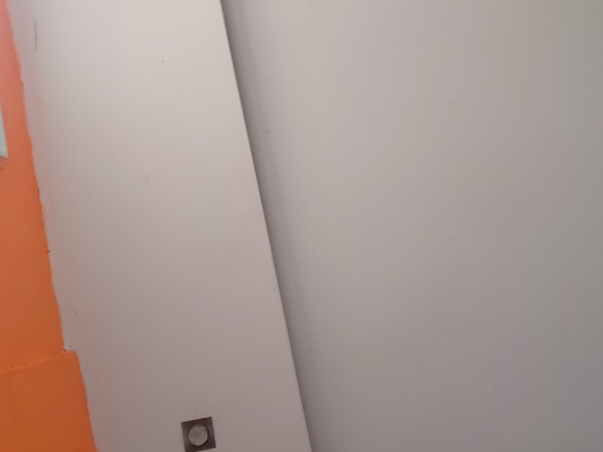 The image captures an indoor architectural detail, primarily featuring two large, smooth, light-colored wall surfaces that meet at a sharp diagonal line, creating a sense of depth or an angled corner. A vibrant, solid orange-painted section of a wall is visible along the left edge of the frame, contrasting with the lighter main surfaces. Near the bottom left, positioned at the junction of the orange and light-colored walls, is a small, square, metallic-framed object with a circular, slightly recessed or translucent center, likely a flush-mounted light fixture, a switch, or an electrical outlet cover. The lighting is even and diffused, without strong shadows, suggesting an interior space. No people, discernible activities, or text are visible. The scene reflects a minimalist aesthetic with distinct color blocking and clean lines. This interior detail is located in Santa Ana, Venezuela.