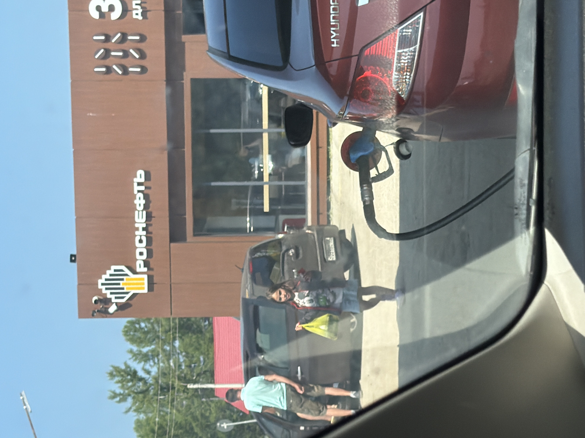 In Turka, Russia, at a Rosneft gas station, a dark-colored minivan is parked with its driver's side door open. A man in a light blue t-shirt and patterned shorts stands by the open door. A young girl in a blue skirt and patterned jacket is walking away from the minivan, holding a yellow plastic bag. In the foreground, a red Hyundai sedan is being refueled, with a gas pump nozzle inserted into its fuel port. The gas station building is brown with prominent "Rosneft" signage. The sky is clear and blue, and trees are visible in the background.