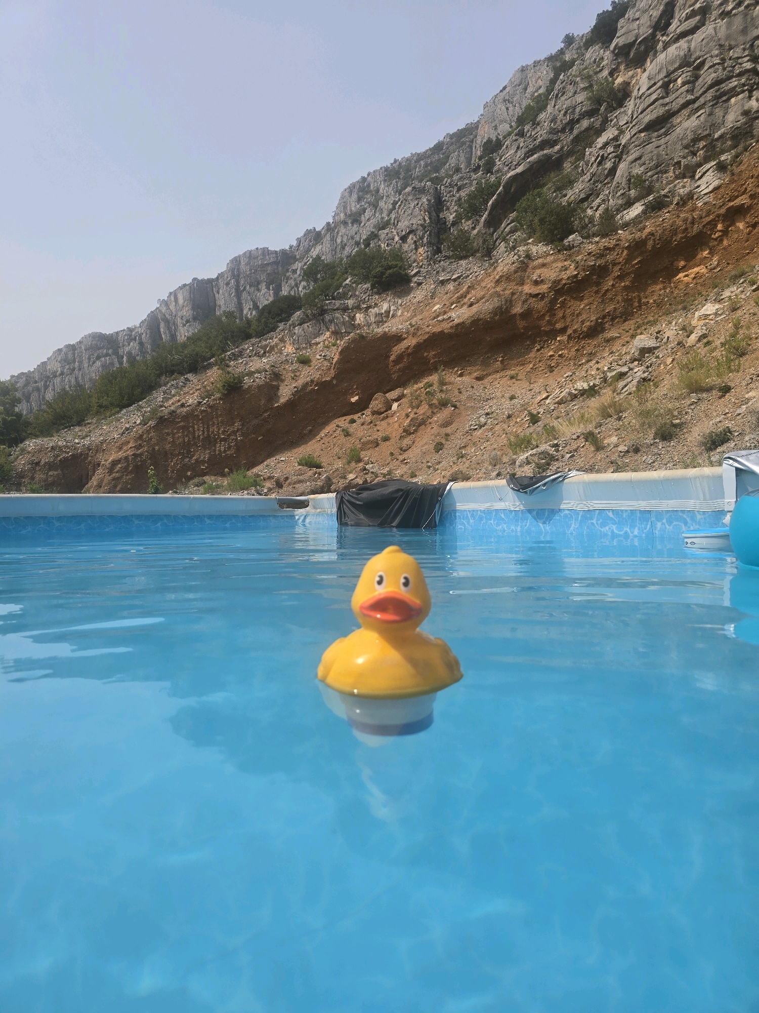 A yellow rubber duck toy floats in the bright blue water of an in-ground swimming pool. The pool is situated at the base of a steep, rocky mountain range. The mountain is covered in sparse vegetation, with a large section of exposed, reddish-brown earth higher up. The sky above is clear and a pale blue. There is a black tarp partially covering the pool on the right side, and a blue inflatable object is visible at the far right edge of the pool. The scene appears to be outdoors in a natural, mountainous environment near Podgorica, Montenegro.
