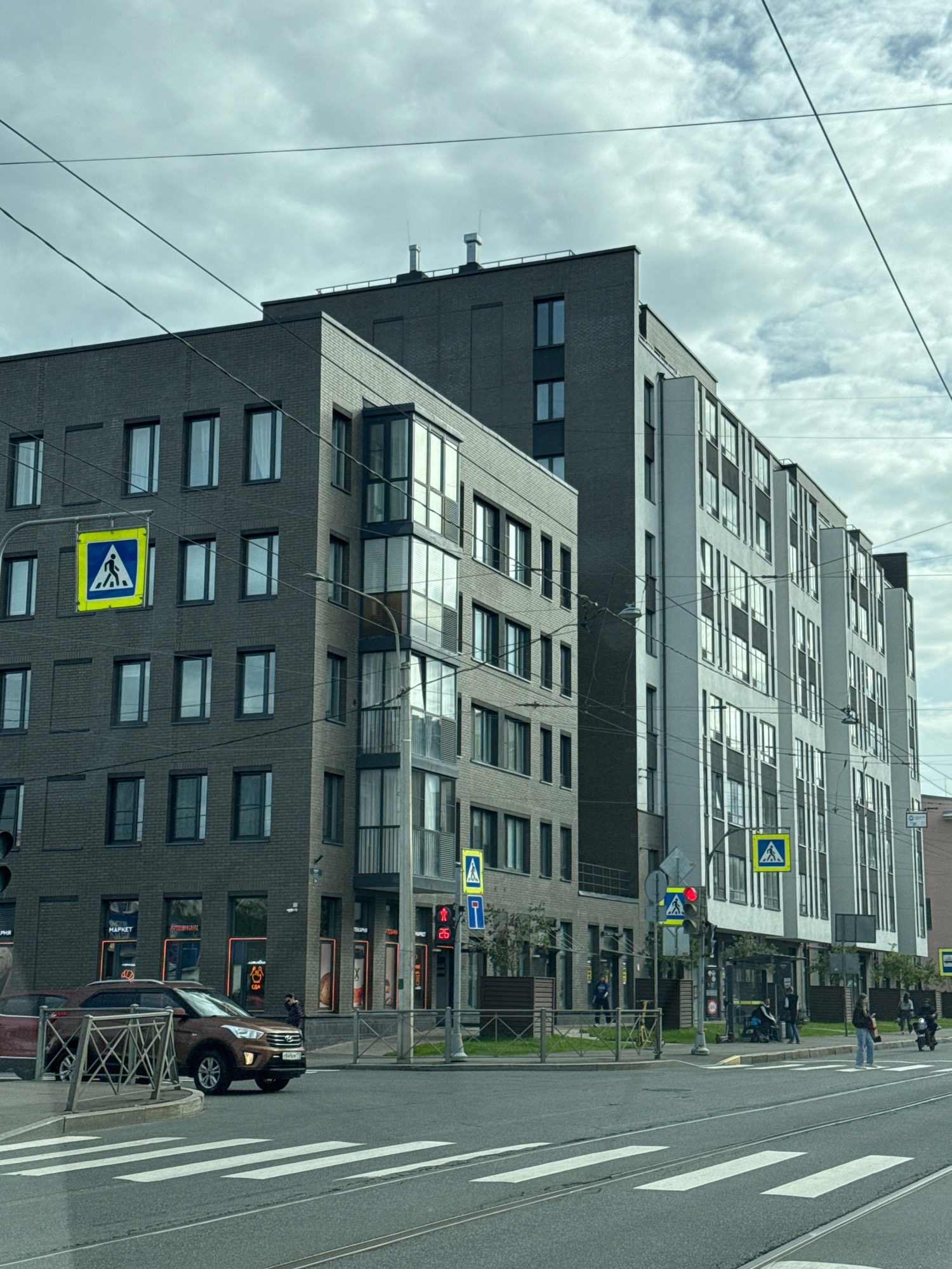 The image depicts a street scene in Saint Petersburg, Russia, featuring modern multi-story buildings with a mix of dark brick and white facades. Several pedestrian crossing signs are visible, along with traffic lights, including one showing a red signal. A brown SUV is stopped at the intersection, indicating a pause in traffic. Pedestrians are present on the sidewalks and one person is crossing the street. The sky is overcast with clouds. Overhead power lines are a prominent feature of the urban landscape. The ground is paved with asphalt, and there are tram tracks on the road.