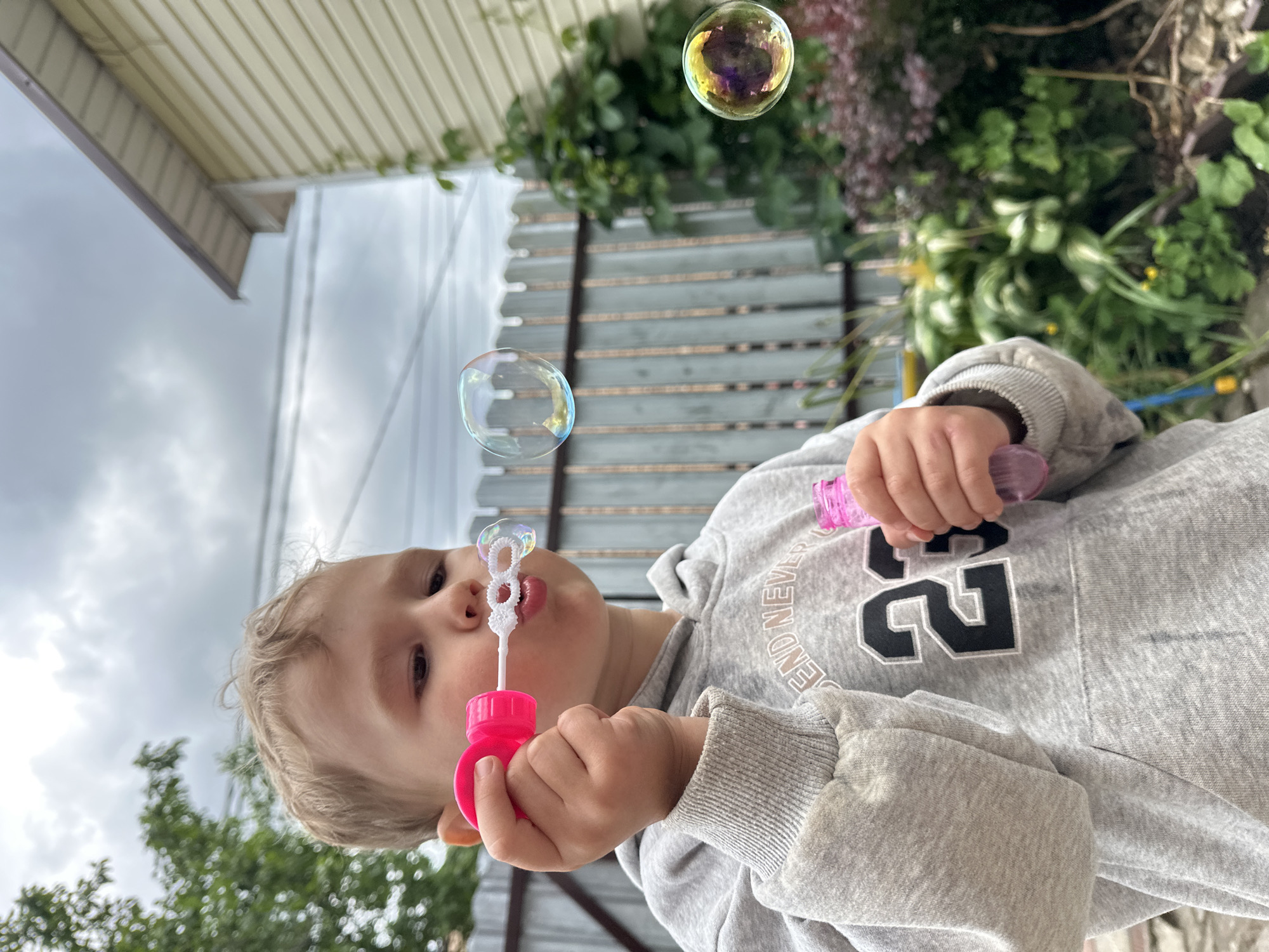 A young child is blowing bubbles outdoors in what appears to be a backyard. The child is wearing a grey hoodie with the number "23" prominently displayed on the front. They are holding a pink bubble wand with a circular blowing apparatus and a small pink bottle containing bubble solution. Several iridescent bubbles are visible in the air. In the background, a wooden fence, green foliage, and a light-colored building with a beige, horizontal siding are present. The sky is overcast. The location is identified as Nizhny Novgorod, Russia.