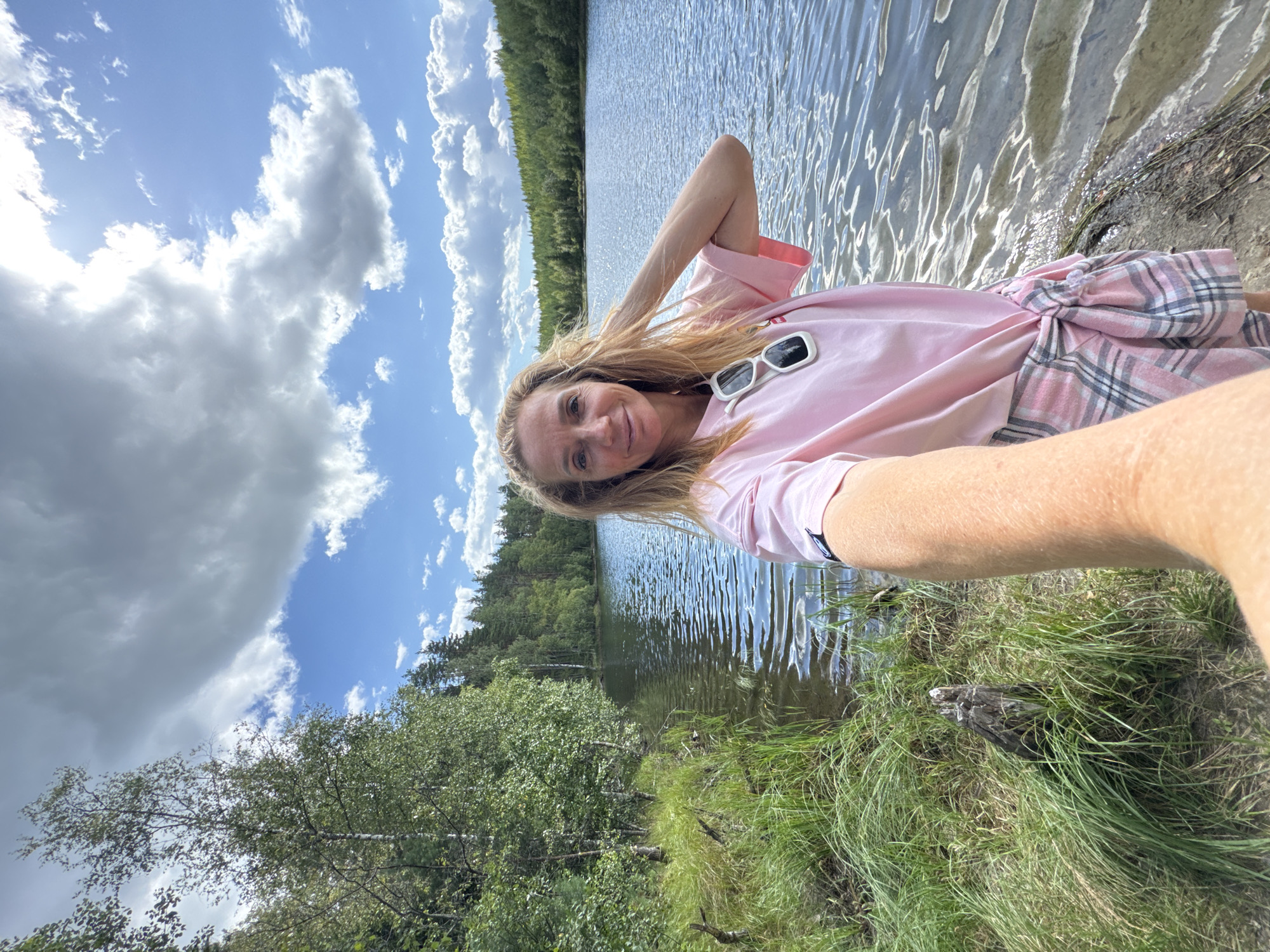 In Lumivaara, Russia, a woman with blonde hair and sunglasses perched on her shirt takes a selfie by a lake. The lake is surrounded by trees and the sky is cloudy. She is wearing a pink t-shirt and a pink plaid skirt. The water is calm with ripples, and there is grass and a tree stump on the shore.
