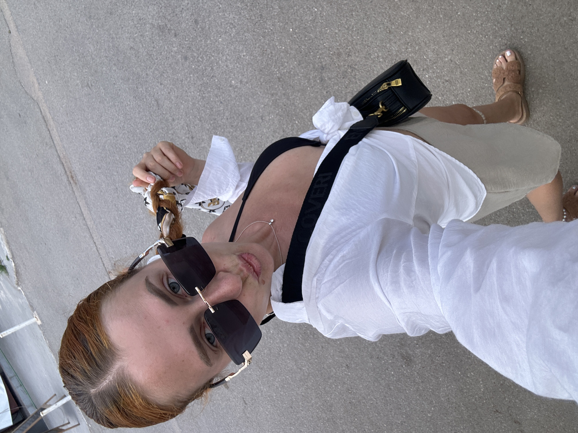 A Caucasian woman with auburn hair, wearing sunglasses, a black bikini top, and a white open button-down shirt, poses for a selfie on a paved street in Budva, Montenegro. She holds a patterned scarf in her right hand near her face, with her hair braided into a ponytail. She is also wearing a small silver necklace and carrying a black crossbody bag. A light-colored skirt is visible around her waist, and she is wearing tan fuzzy slides on her feet. The background shows a grey asphalt surface with white road markings.