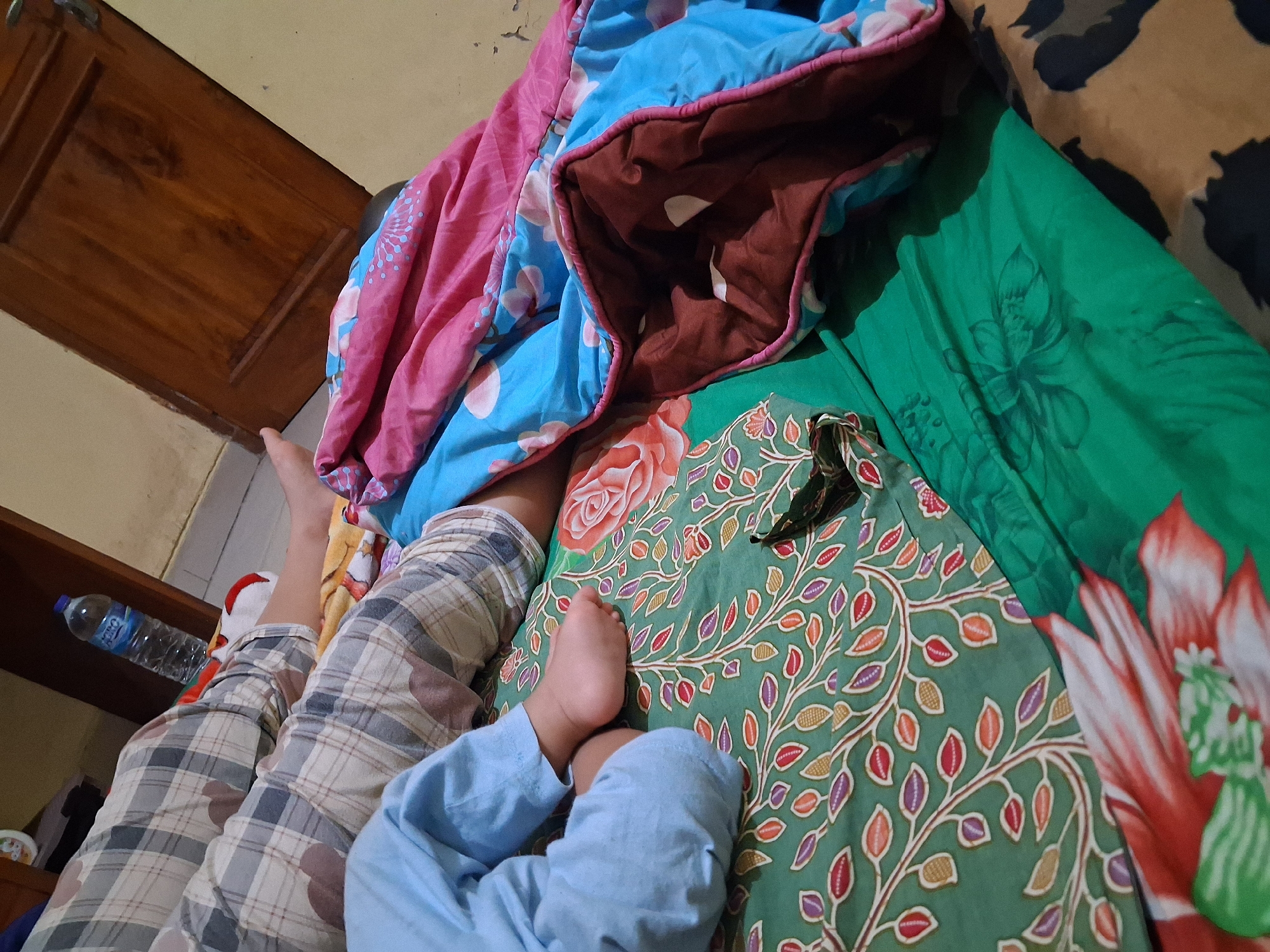 The image shows the lower half of two individuals lying on a bed. One person is wearing plaid pajama pants, and the other is wearing blue pants. A baby's foot is visible, positioned on a green batik-patterned sheet. The bed is covered with colorful blankets, one with a pink and blue floral design and another with a brown underside. A plastic bottle of water is on a nightstand in the background, next to a wooden door. The room appears to be a bedroom in a home, possibly in Pendawa, Indonesia.