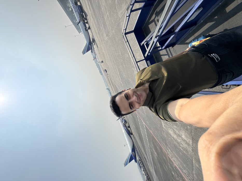 A man poses for a selfie at an airport tarmac. He wears a green shirt and dark jeans with a silver belt buckle. Behind him, several indigo-colored airplanes are parked in a row. The tarmac is gray and marked with white lines. Part of a blue metal railing or structure is visible on the right side of the frame. The sky is bright and hazy.