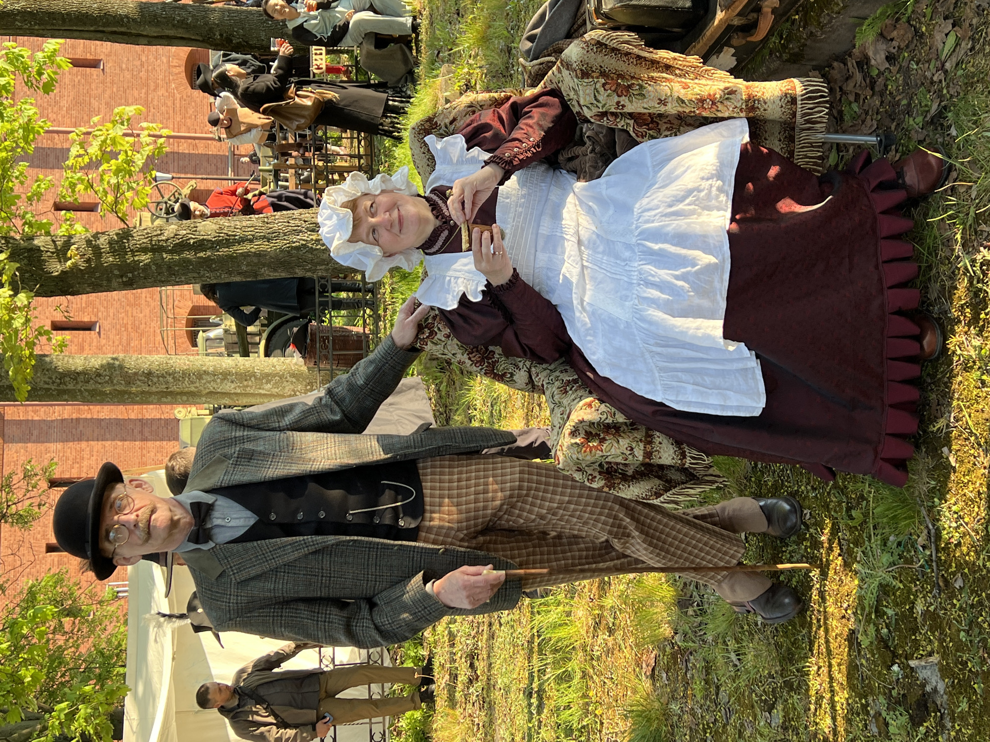 An outdoor scene depicts two individuals in period attire amidst a group of people. A man stands wearing a bowler hat, glasses, a gray tweed jacket, a black vest, a gray collared shirt with a bow tie, and plaid pants. He is holding a thin wooden cane. A woman, wearing a white bonnet and a white apron over a dark red dress, is seated on an ornate chair with a patterned fabric. She is holding a small, dark object in her hand. The setting appears to be in a grassy area near a brick building, possibly in Saint Petersburg, Russia. Other people are seated and standing in the background.