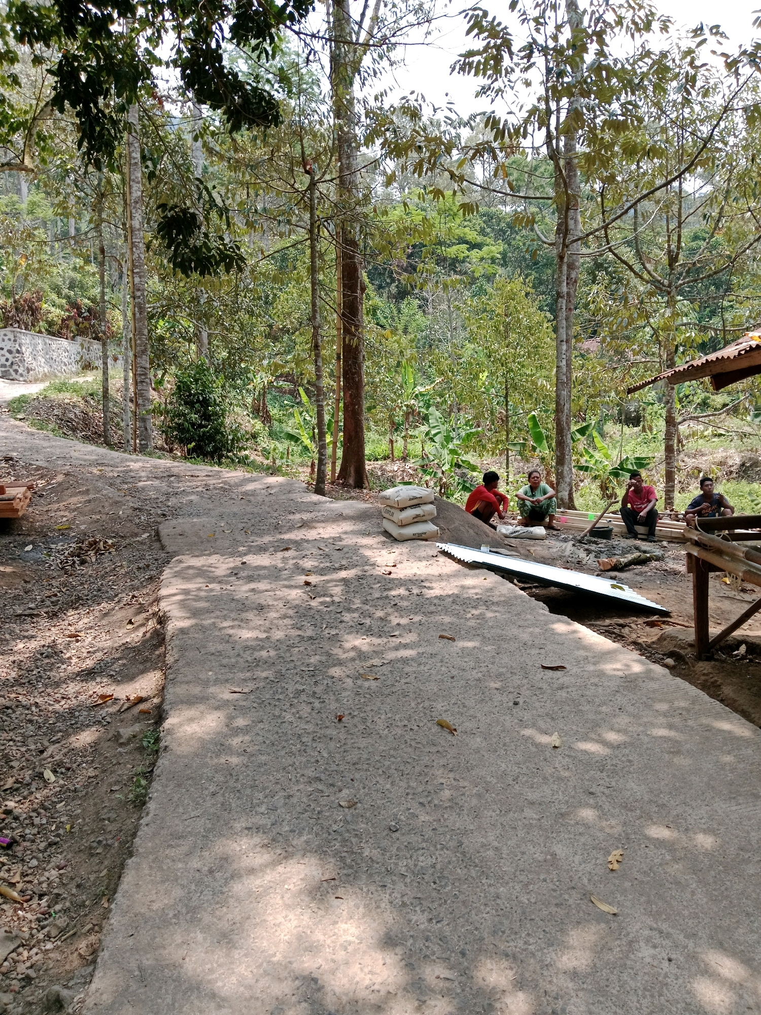 The image depicts a scene outdoors in a rural area, potentially Leuwikoja, Indonesia, characterized by lush vegetation and trees. A rough concrete pathway dominates the foreground, with a slight incline to the left. On the right side of the pathway, a group of four individuals are gathered. Two men in red shirts, one man in a dark shirt, and one woman in a green shirt are seated. To their left, a stack of four large sacks, likely containing building materials like cement, is visible. Behind the people, a collection of wooden planks is stacked, suggesting construction or repair work. A shovel rests on the ground near the planks. A simple, open-sided shelter with a corrugated metal roof is partially visible on the far right. The environment is natural, with trees and foliage surrounding the area, and sunlight filtering through the leaves, casting dappled shadows on the ground.
