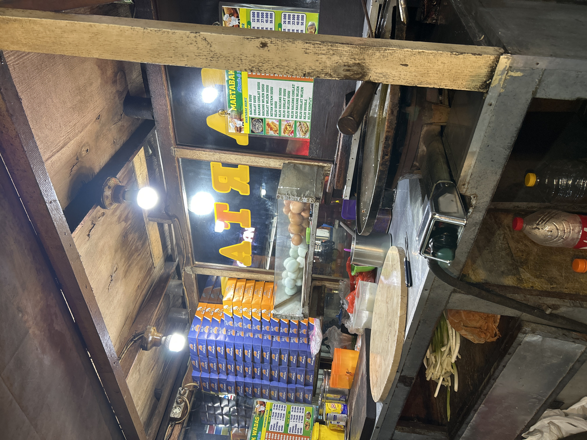 A food stall is pictured at street level in Depok, Indonesia, with a focus on the preparation area for martabak, a savory or sweet pancake. The stall appears to be constructed from wood and metal, with a wooden ceiling that has exposed wiring and lighting fixtures. Behind the counter, a large window displays stacks of blue and orange boxes, possibly containing ingredients or packaging. A menu board is visible on the right side of the window, listing martabak variations with prices.
On the counter, there are several key items: a large round wooden cutting board, a griddle or flat-top cooking surface, and various containers and utensils. A basket of green onions and a loaf of bread are visible in the lower section of the stall, suggesting ingredients being stored. In the background, a display of white and brown eggs is partially obscured. The overall impression is of a busy, functional food preparation environment at a local establishment in Depok.