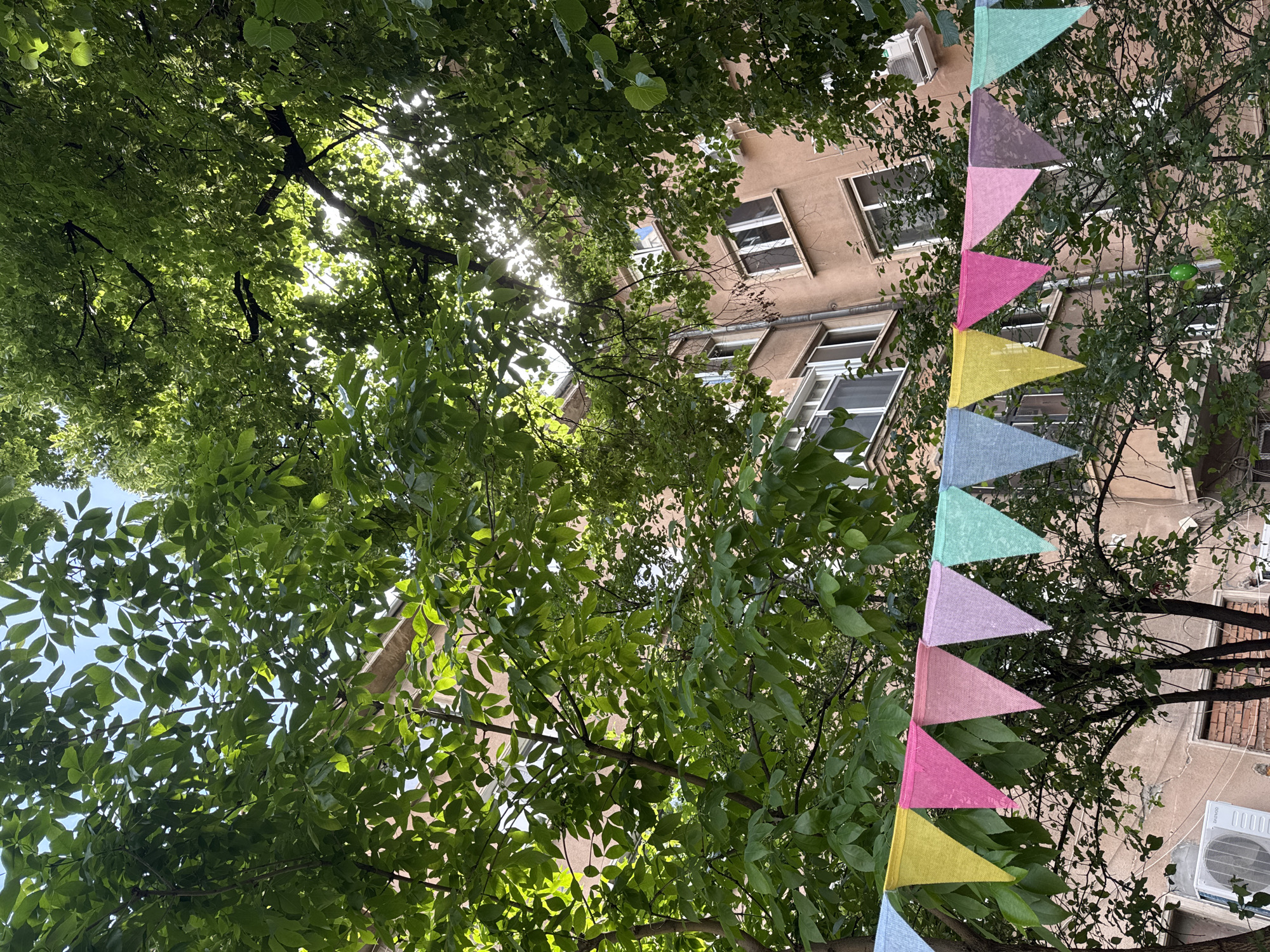 The image depicts an outdoor scene in Sofia, Bulgaria. A building with several windows is visible through dense green foliage. A string of colorful triangular flags hangs diagonally across the right side of the frame. An air conditioning unit is mounted on the building's exterior wall. Sunlight filters through the leaves, creating a dappled effect.