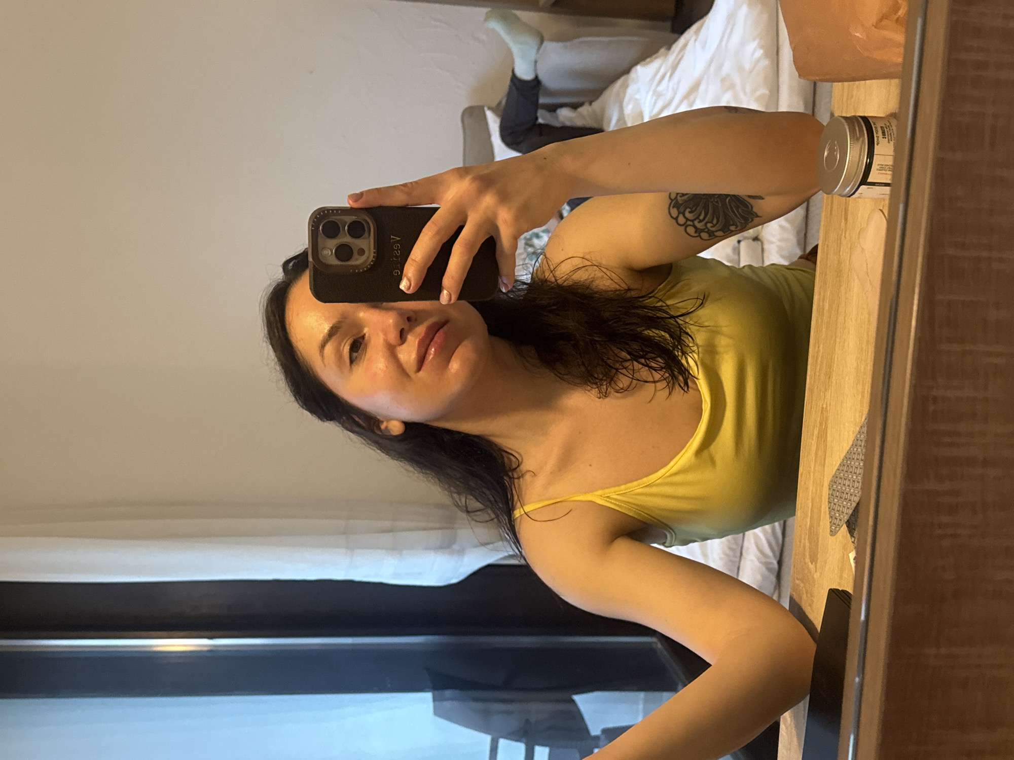 A woman is taking a selfie in a mirror. She is wearing a yellow tank top. Her hair is dark and curly. She has a tattoo on her left arm. The mirror reflects a bed with white sheets and a person in socks and dark pants lying on it. There is also a window in the reflection. It is not possible to identify the location in the city of Krasnovo, Bulgaria.