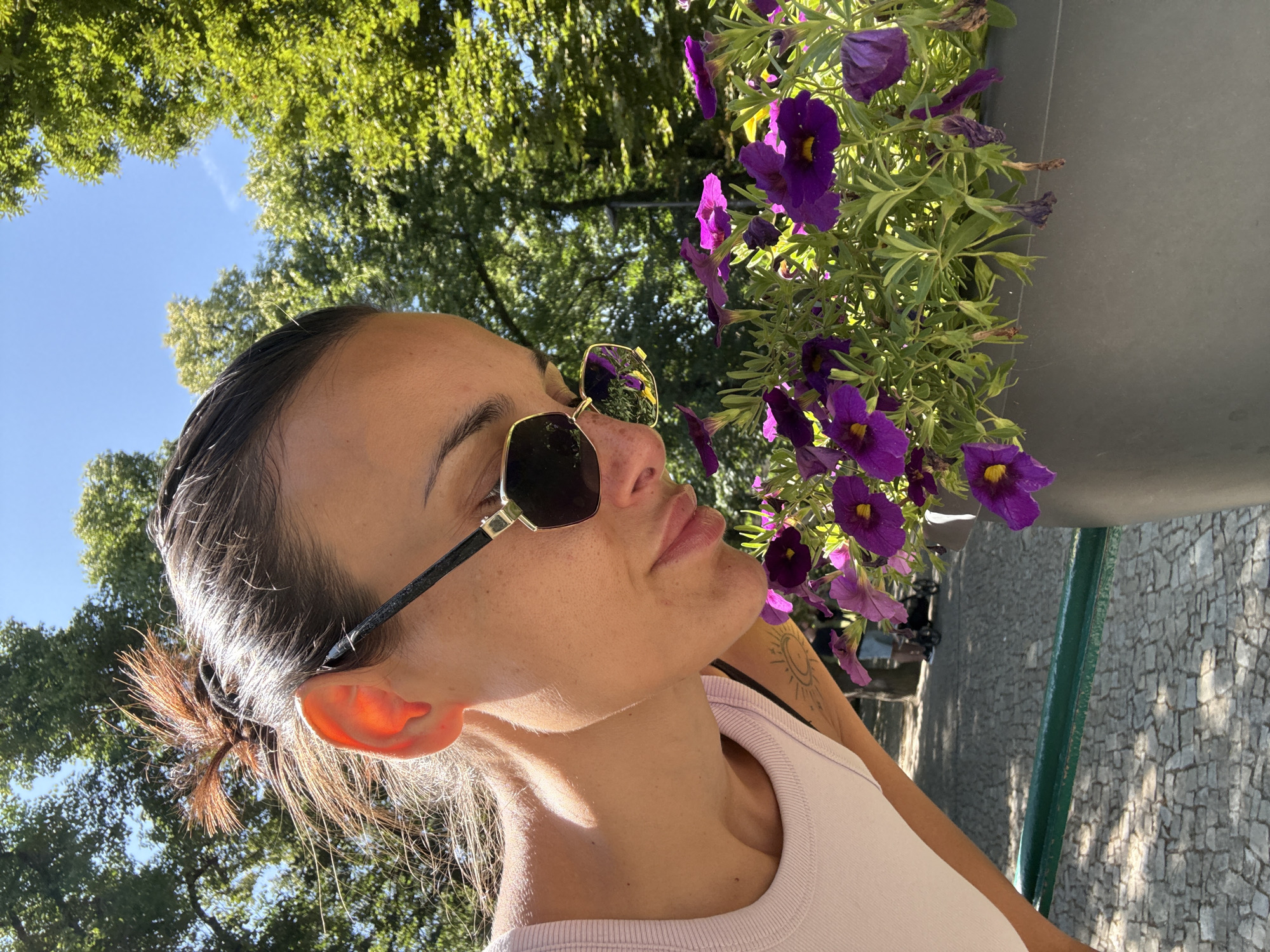 A woman wearing sunglasses and a light pink tank top is leaning down to smell purple flowers in a planter. The woman has her hair pulled back in a bun. She has a tattoo of a sun on her left arm. The planter is gray. In the background, there are trees and a cobblestone path. The scene appears to be outdoors in a park or garden. The image does not contain enough information to determine the specific location in Prague, Czechia.