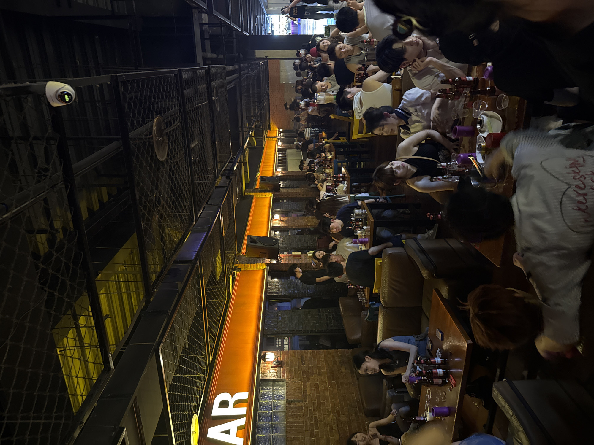 The image depicts a busy, open-air bar in Liangjiang, China. The bar, signified by a large "BAR" sign, is crowded with people sitting at tables. Most of the patrons appear to be young adults. Tables are filled with beer bottles and glasses, suggesting a lively atmosphere. The location appears to be within an urban setting. Metal scaffolding and netting are visible above, and the overall lighting is low, creating an ambiance. A security camera is mounted on the structure above. Brick walls and seating areas with sofas line the space.