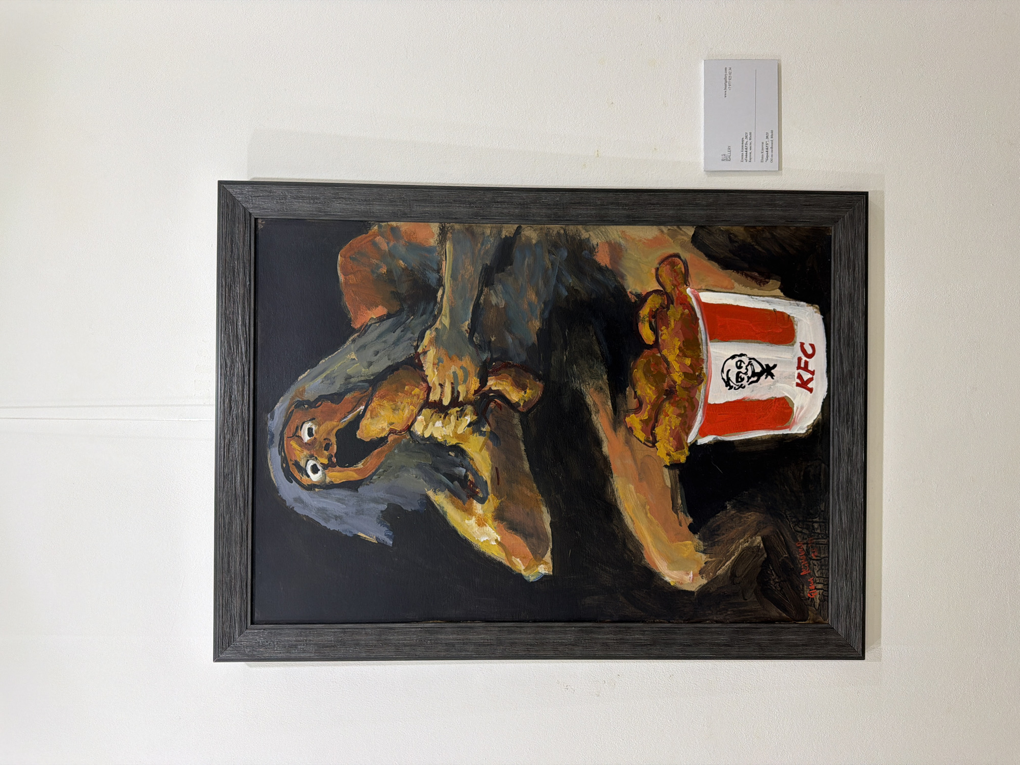 A framed painting is displayed horizontally on a plain white wall, likely within an art gallery or exhibition setting in Moscow, Russia. The painting features two highly stylized, almost grotesque human figures rendered with broad, visible brushstrokes in shades of orange, brown, grey, and dark blue against a dark background. On the left, a larger, dominant figure, possibly an old woman or witch-like character with long grey hair and piercing white eyes, appears to be holding or interacting with a smaller, infant-like figure. To the right of these figures, a prominent red and white KFC bucket, bearing the brand's logo, is shown filled with what appears to be fried chicken pieces. The artistic style is raw and expressionistic, creating an unsettling visual commentary.
Above and to the right of the painting, a small white informational label is affixed to the wall. Visible text on the label includes "BIG GALLERY", the artist's name "Elena Kinovar", and the painting's title and year: "«Fond KFS», 2023". The label also provides the gallery's website "www.hisartgallery.com" and a phone number "+7 977 925 02 34". The "KFC" logo is clearly visible on the bucket within the painting itself.