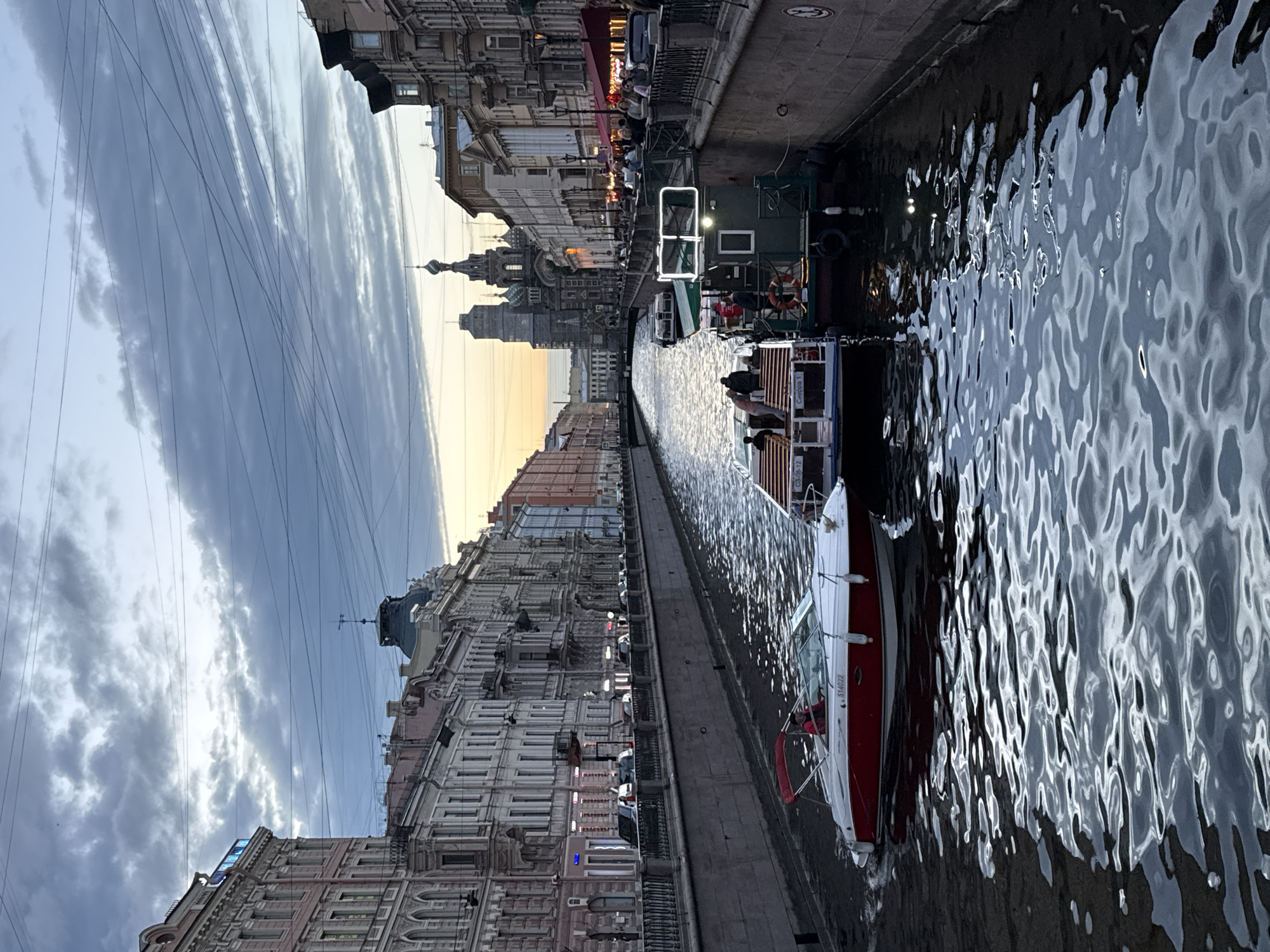 A wide canal runs through a European-style city, bordered by ornate, multi-story buildings with numerous windows and decorative facades. Several boats navigate the canal. In the foreground, a red and white speed boat with a canopy is moving, with one person visible at the helm. Behind it, a larger, green boat, likely a tour boat, has several passengers visible on its deck. Further in the background, the iconic onion domes of the Church of the Savior on Spilled Blood are visible, silhouetted against a cloudy sky with a hint of sunset light peeking through. Overhead, a complex network of electrical wires crisscrosses the sky. The water in the canal is dark and reflects the sky and surrounding structures, creating rippling patterns. The overall scene depicts a canal tour in St. Petersburg, Russia, likely during the evening.