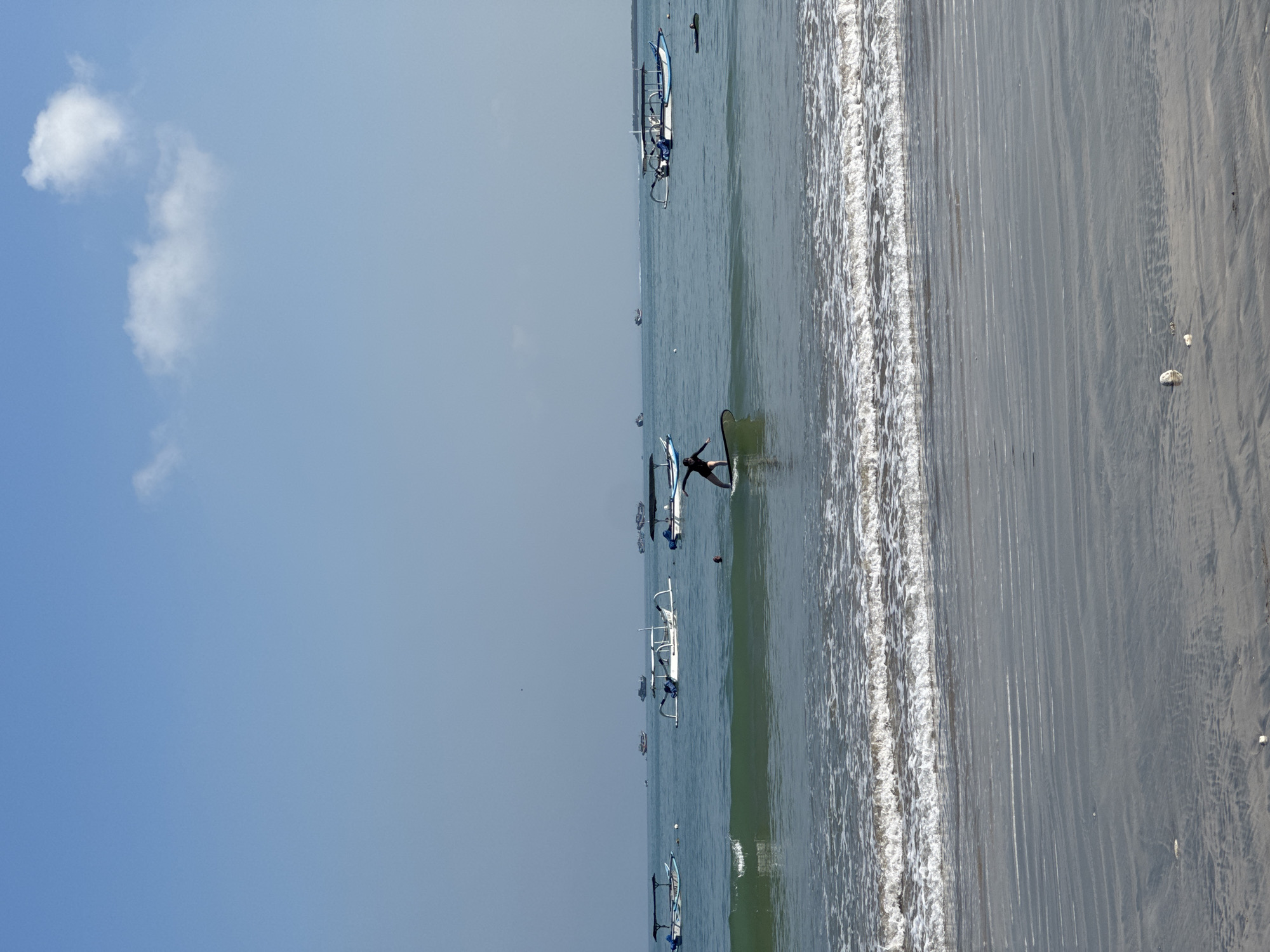 This image depicts a beach scene in Kuta Selatan, Indonesia. The beach is visible on the right side of the frame, with wet sand transitioning into drier sand. Waves are crashing along the shoreline. In the ocean, there are several traditional outrigger boats (Jukung) with blue and white detailing. A person, likely a surfer, is lying on a surfboard in the water near one of the boats. The sky is clear and blue with a few clouds on the left side of the image.
