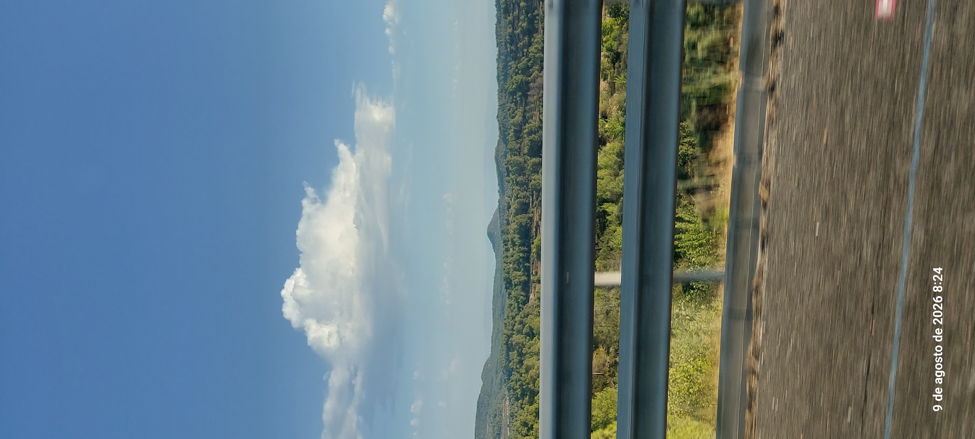 The image shows a view from a moving vehicle, looking towards a landscape with rolling hills covered in dense green trees. A bright white cloud is prominent in the clear blue sky. In the foreground, a silver metal guardrail runs horizontally across the frame, partially obscuring the view of the road ahead which appears blurred due to motion. The lower right corner of the image has text overlayed, reading "9 de agosto de 2026 8:24", which indicates the date and time the photo was taken. The text suggests the location is Vinaixa, Spain, implying the scene is a road in or near that municipality on August 9, 2026, at 8:24 AM. There are no people or animals visible in the frame. The environment is natural, with a focus on the expansive sky and the tree-covered terrain.
