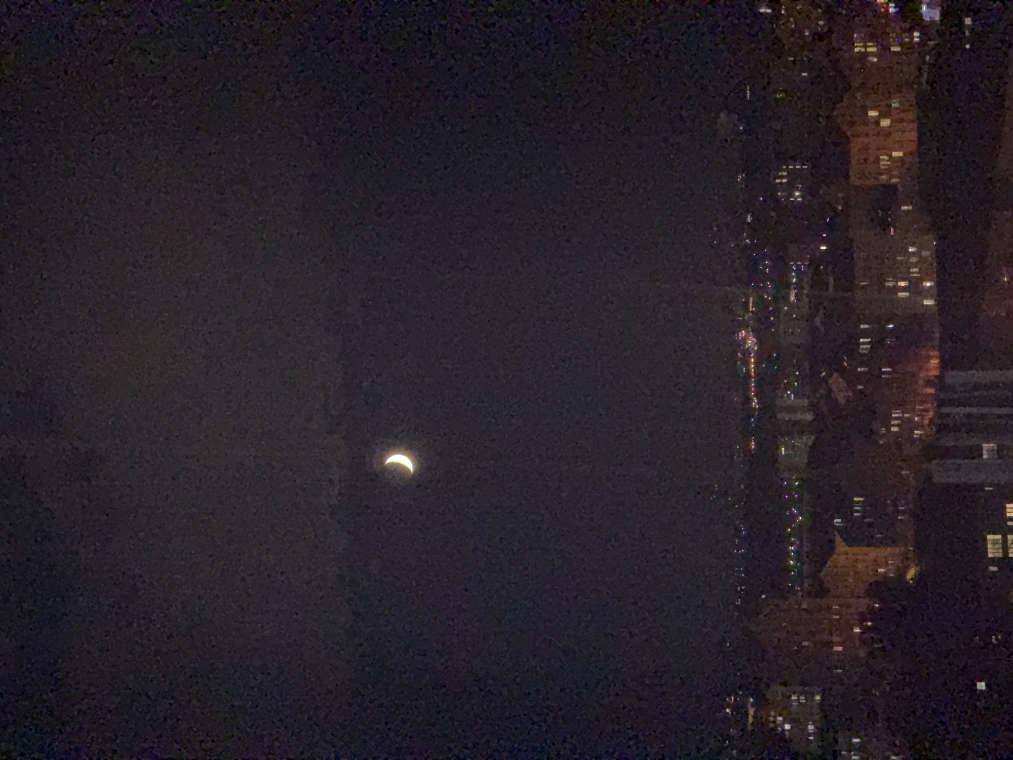 A crescent moon is visible in the night sky above a city skyline. The moon is illuminated and appears bright against the dark, cloudy sky. Below the moon, the lights of buildings and streetlights can be seen in the distance, indicating an urban environment. The city appears to be Kyiv, Ukraine, as suggested by the user's prompt. The buildings in the foreground are dark, with some windows illuminated, suggesting residential or commercial structures. The overall scene depicts a nocturnal view of a city with the moon as a prominent celestial object.