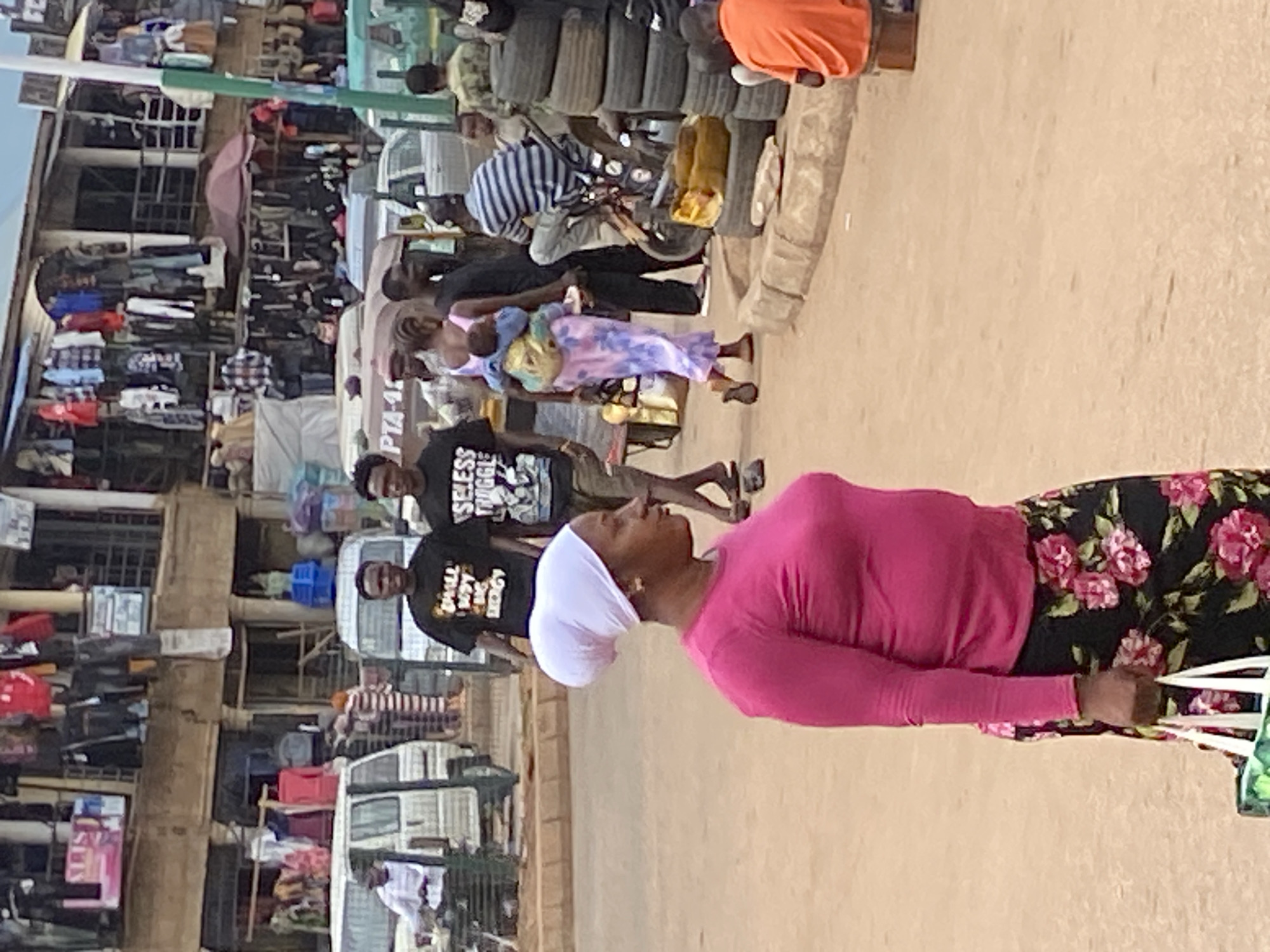 This image captures a vibrant and busy street market scene in Bukuru, Nigeria, during the daytime. The perspective is from the street level, looking towards a row of market stalls and buildings.
In the foreground, on the right side of the frame, a woman is seen from the chest up, facing right. She is wearing a bright pink long-sleeved top and a black skirt with a vibrant pink and green floral pattern. She has a white head covering and appears to be holding white plastic bags. Her expression is neutral as she looks forward.
Behind her, slightly to the left, are two men. The man closer to the foreground is wearing a black t-shirt with white text that reads "SHALL NOT BE MOVED". The man further back is also wearing a black t-shirt with white text, which appears to say "BLESS THUGGZ". Both men are looking in the same direction as the woman.
Further into the midground, the street is bustling with people and activity. A woman in a light purple patterned dress is seen carrying a baby on her back, walking among other pedestrians. To her left, a person is seated on a motorcycle, surrounded by stacks of tires, indicating a tire vendor. Another individual is visible wearing a blue and white striped shirt.
On the left side of the frame, a row of market stalls and multi-story buildings lines the street. These structures are packed with various goods on display, including clothing (shirts, trousers, jackets) and bags, suggesting a general merchandise market. An open, reddish-brown umbrella is visible within one of the stalls. Several vehicles, including vans or minibuses, are parked or moving along the street. One vehicle clearly shows "PTA-14" on its side. The ground appears to be a light-colored, possibly paved, surface. The bright lighting suggests a sunny day. The overall scene conveys a dynamic and lively urban market atmosphere.