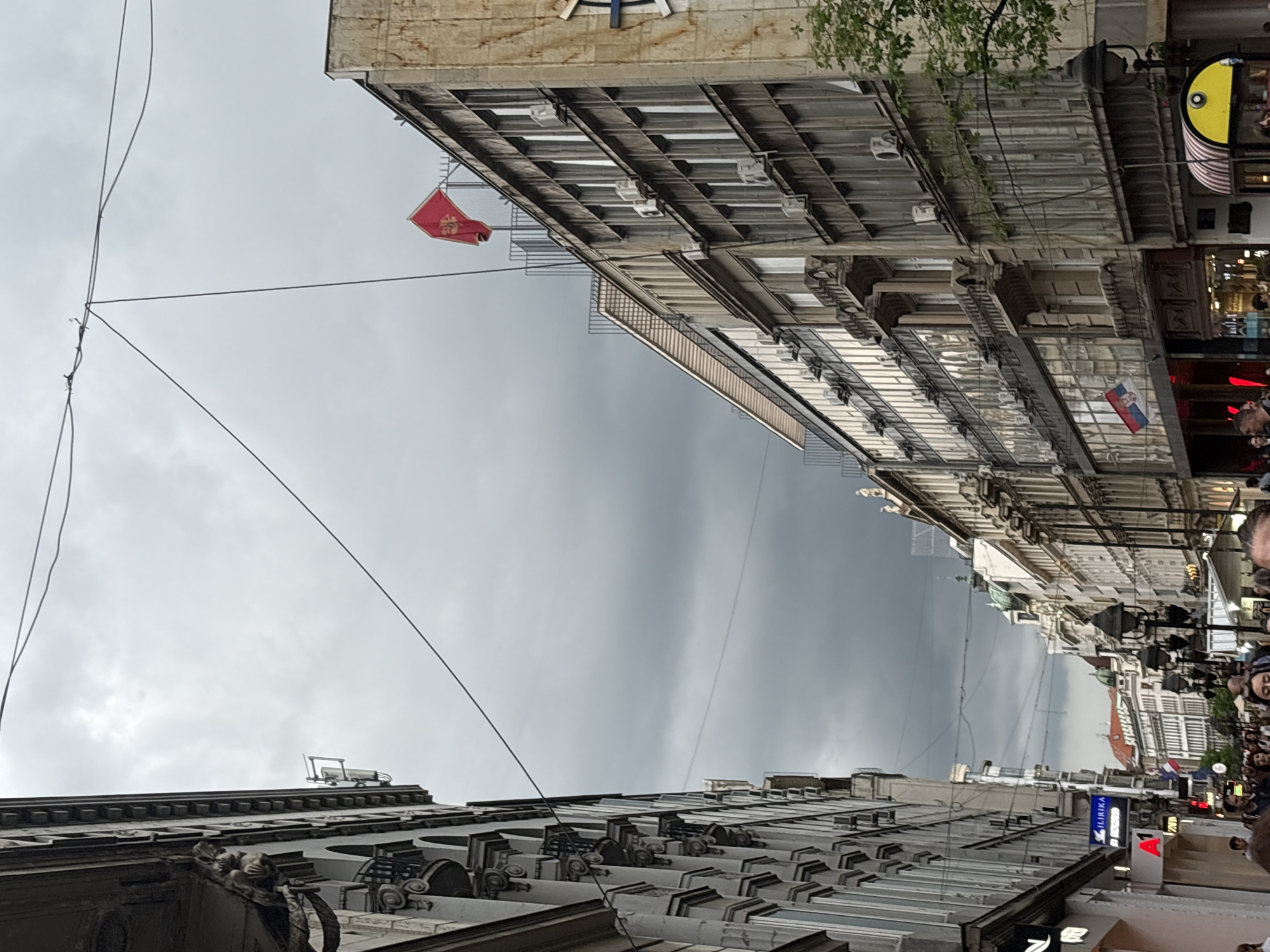 The image depicts a street scene in Belgrade, Serbia, taken from a low-angle perspective, looking upwards towards buildings and a cloudy sky. Two ornate buildings frame the view. Power lines crisscross the sky. A red flag with a symbol is attached to one building. Further down the street, flags of Serbia are visible. The street is crowded with people. Signage and shops are present along the street level.