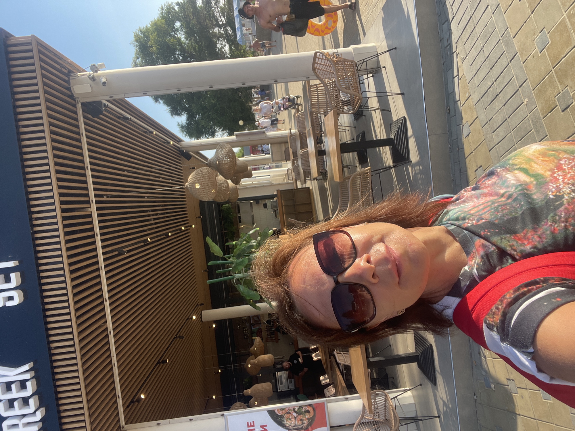 The media file is a selfie taken outdoors on a sunny day. The subject is a woman with auburn hair, wearing sunglasses and a colorful patterned top and a red jacket. She appears to be smiling slightly.
In the background, the scene depicts an outdoor dining area with several tables and chairs made of wicker. The setting appears to be a modern establishment with a facade featuring horizontal wooden slats. Several decorative lamps with a woven texture hang from the ceiling.
Further in the background, there are glimpses of other people, some appearing to be near a pool with a man holding a yellow inflatable ring. Trees are visible, suggesting a pleasant outdoor environment. The ground is paved with rectangular stones.
The time of day appears to be daytime due to the bright sunlight and shadows. The overall atmosphere seems relaxed and summery.
Visible text includes the word "CREEK" in large white letters on a dark blue background on the left side of the image. There is also partial text visible on a sign below that, which appears to be a food advertisement with an image of seafood.