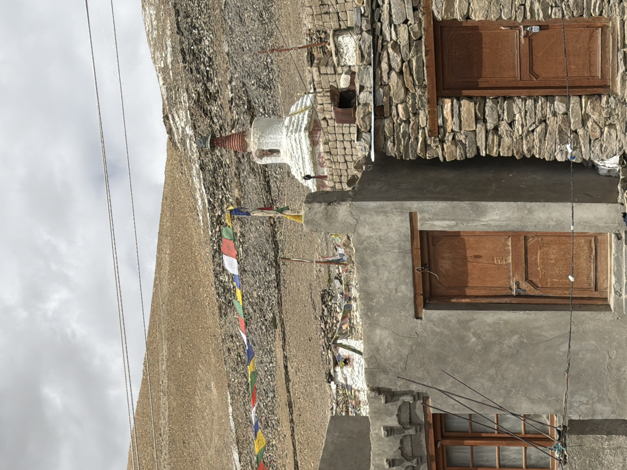 The image shows a dry, mountainous landscape near Karzok, India, with a cloudy sky. In the foreground, there are two buildings. The building on the left is made of grey concrete with a wooden-framed window. A wooden door is visible to its right, set within a stone wall. The building on the right is constructed entirely of stacked stones and features a wooden door. Prayer flags in various colors are strung across the scene, adding a touch of vibrancy to the otherwise muted environment. Behind the buildings, a stupa, a Buddhist religious monument, stands prominently. The landscape is characterized by barren, rocky slopes, suggesting a high-altitude, arid region. Power lines are visible stretching across the upper portion of the image.